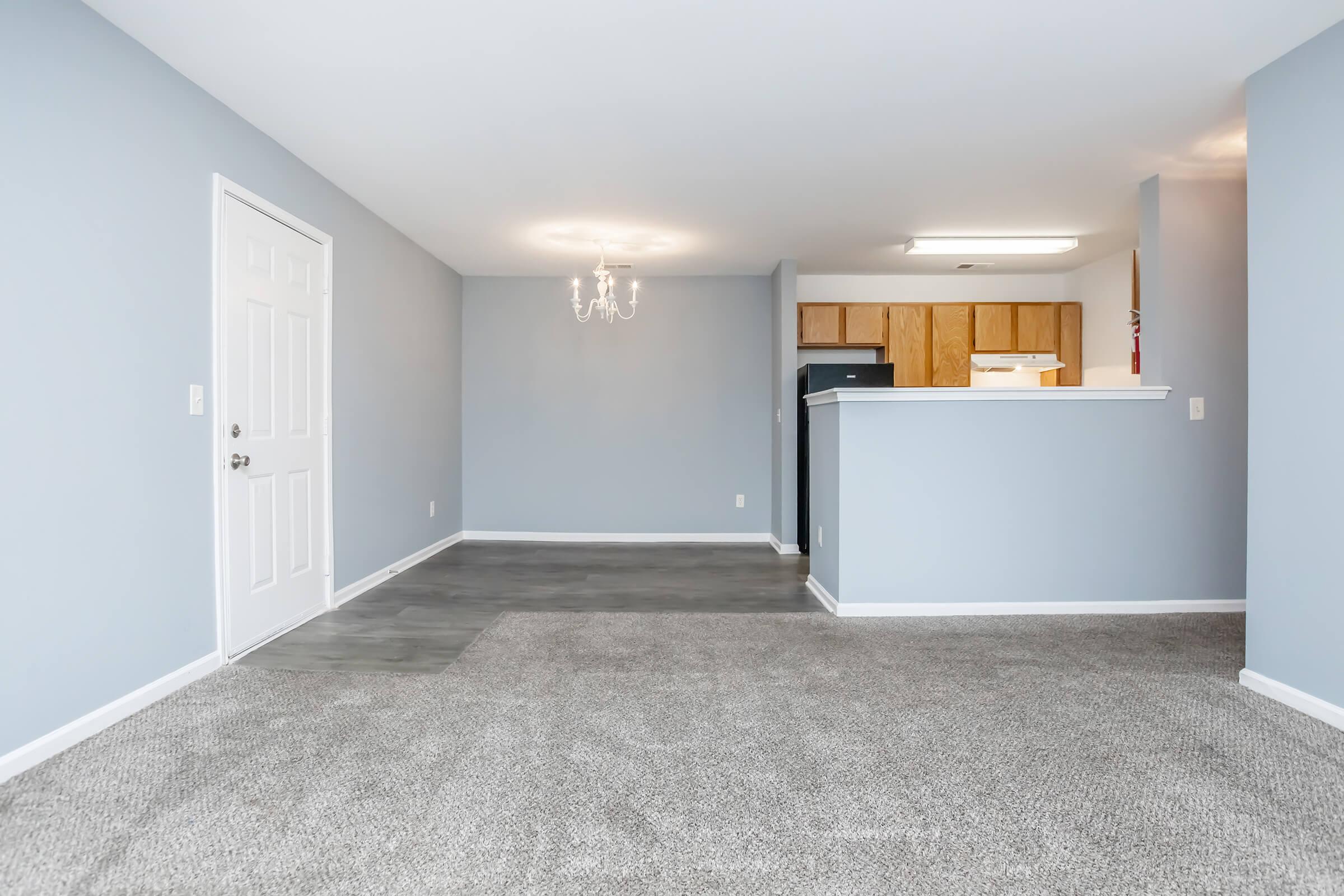 Spacious living area featuring light gray walls and carpeted flooring. A doorway leads to the entrance, while a chandelier hangs from the ceiling. The kitchen area with wooden cabinets is visible in the background, providing an open layout. Natural light illuminates the room, creating a welcoming atmosphere.