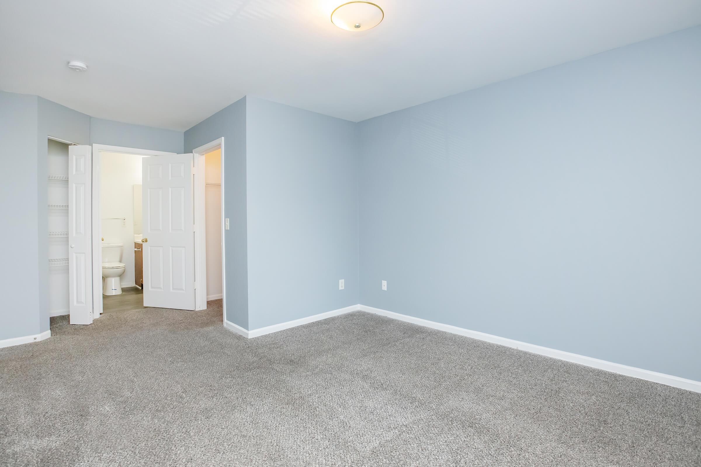 A spacious, light blue room with new carpet and a ceiling light fixture. A doorway leads to a bathroom, partially visible. The walls are undecorated, providing a blank canvas for personalization. Ideal for various uses, such as a bedroom or office space.