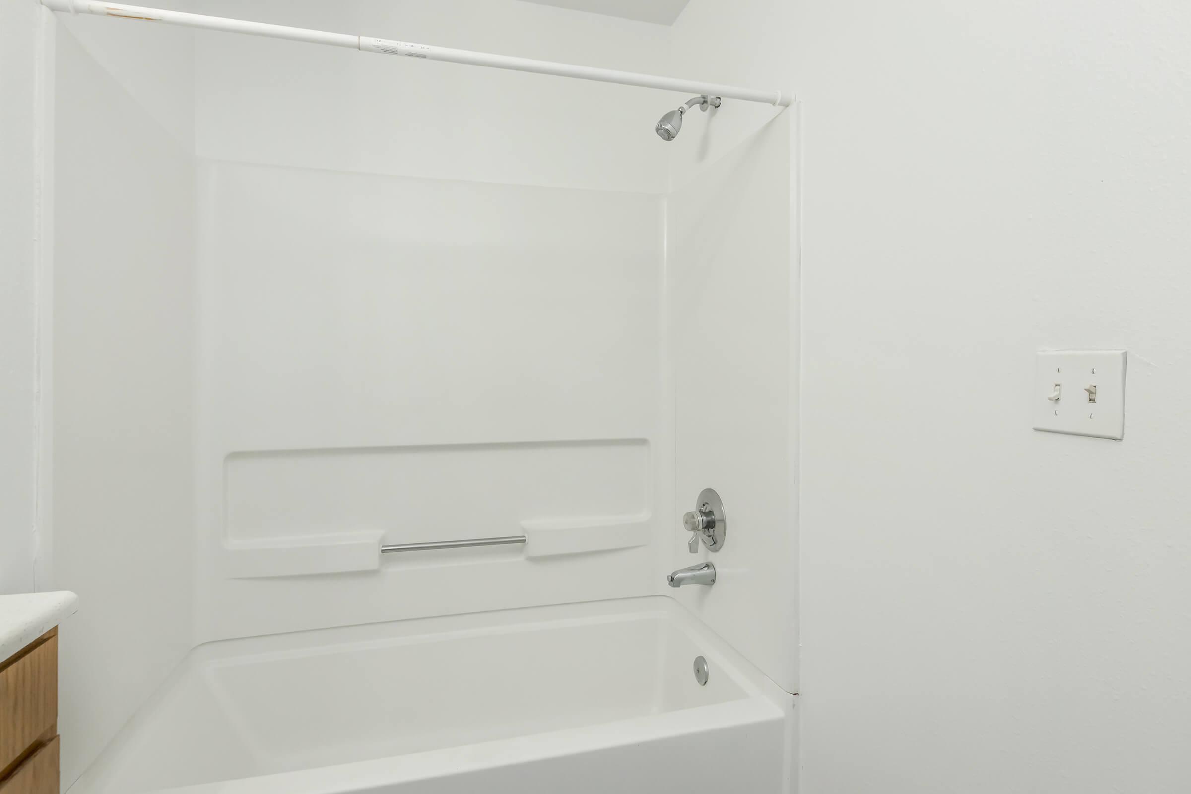 A clean, white bathroom with a simple bathtub and shower combination. The walls and tub are entirely white, creating a minimalist look. There's a silver showerhead and a grab bar installed. A light switch is visible on the right side, and the overall space appears bright and inviting.