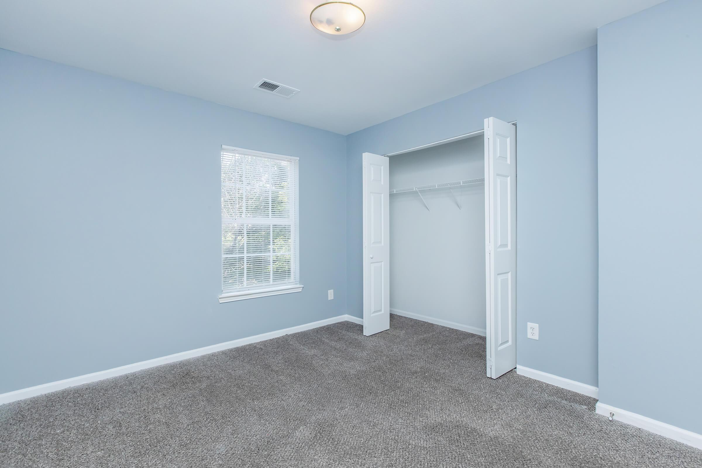 A vacant room with light blue walls and carpeted floor. The space features a window allowing natural light, and an open closet with sliding doors. The ceiling has a simple light fixture, and the room appears clean and well-maintained, suitable for various uses.