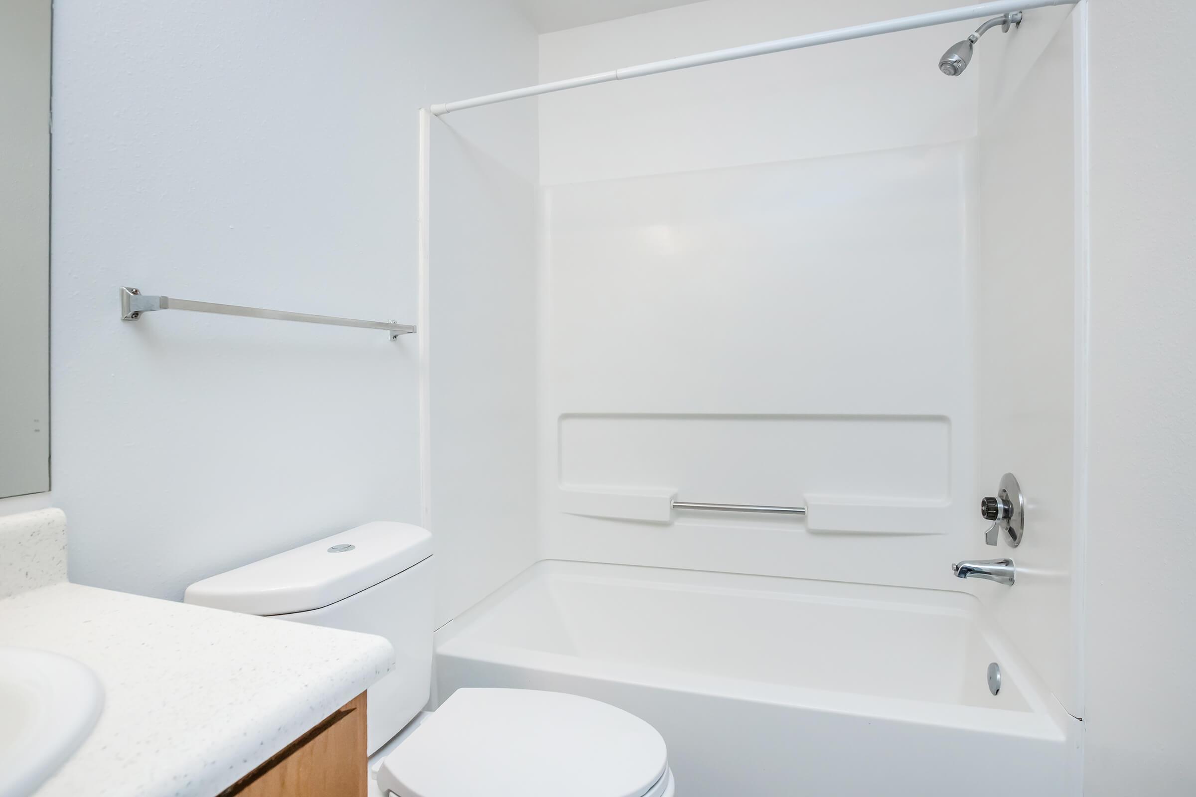A clean and simple bathroom featuring a white bathtub, shower, and toilet. The walls are painted in a light color, and there is a towel rack mounted beside the bathtub. The countertop appears to be a light-colored material, providing a minimalistic and modern aesthetic.