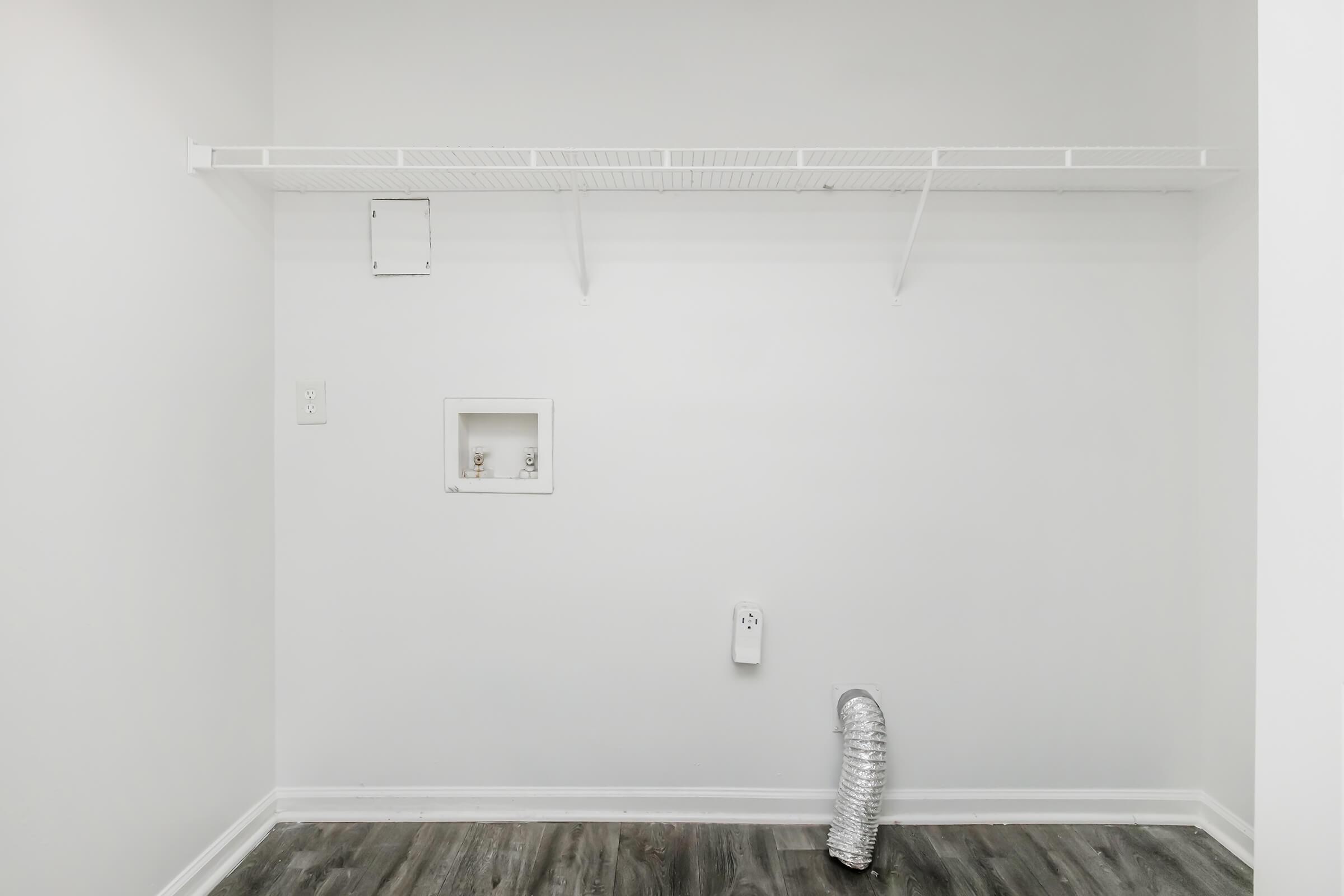 A clean, empty room with white walls and a vinyl floor. There is a wire shelf mounted on the wall, a small access panel, and a vent with insulated ducting on the floor. The space appears ready for storage or installation of appliances.