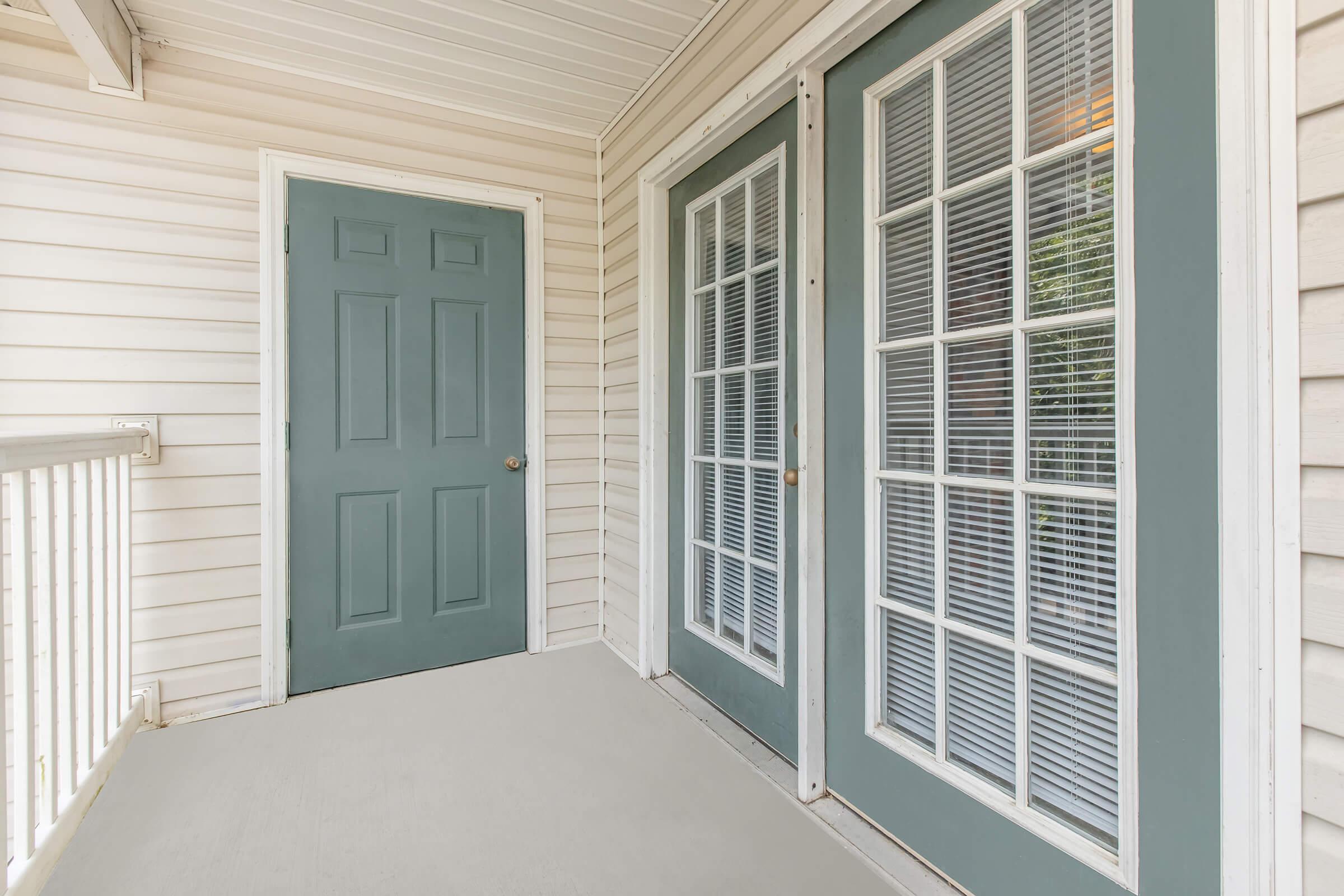 A light-colored porch or entryway featuring a closed blue door and two pairs of double doors with glass panels. The walls are white wood siding, and the floor is a light gray surface. Natural light is visible through the glass doors, creating an inviting atmosphere.