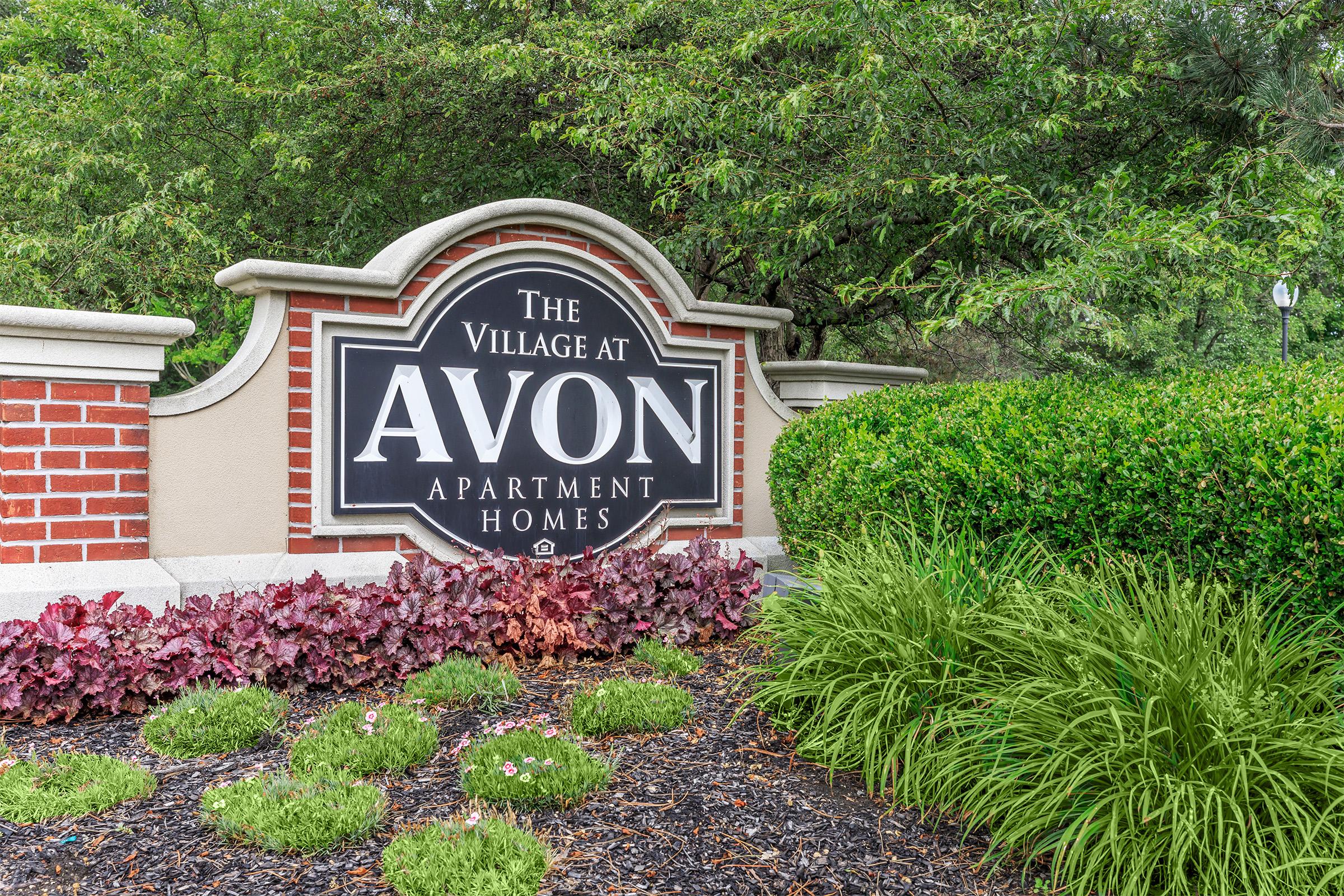 Sign for "The Village at Avon Apartment Homes" surrounded by greenery and colorful landscaping with soil-covered flowerbeds.