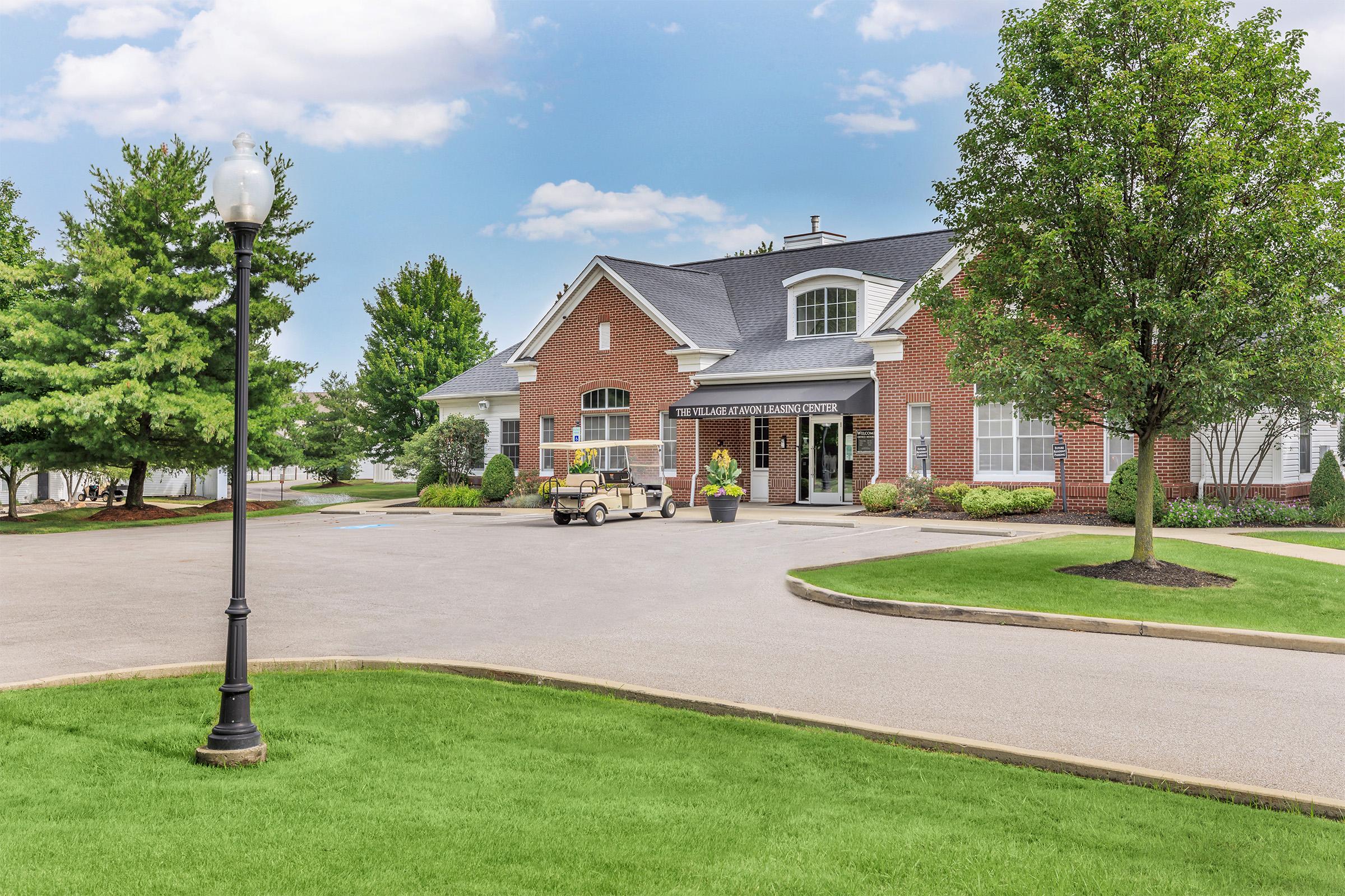 A well-maintained brick building with a gray roof, surrounded by lush green trees and landscaping. There is a circular driveway leading to the entrance, with a golf cart parked nearby. A lamppost stands on the left side, enhancing the inviting atmosphere of the entrance.