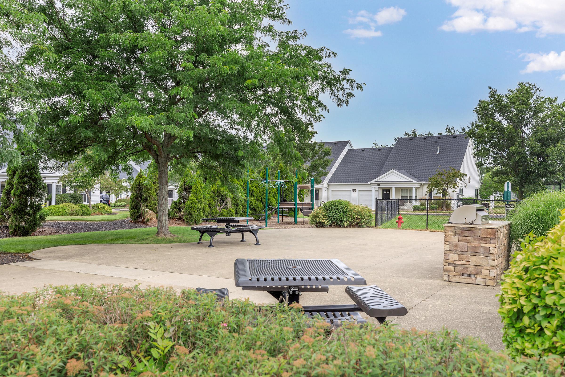 A peaceful outdoor area featuring a concrete patio with picnic tables, surrounded by greenery. In the background, there are residential buildings with black roofs, and a playground structure partially visible. The sky is clear with a few clouds, creating a serene atmosphere.