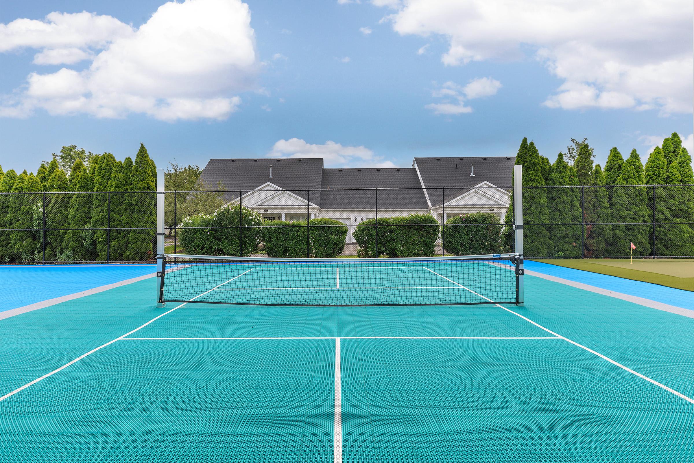 A view of a tennis court featuring a bright blue surface and a net centered in the foreground. In the background, there are lush green hedges and a large, modern house with a gray roof. The sky is partly cloudy, adding a serene atmosphere to the scene.