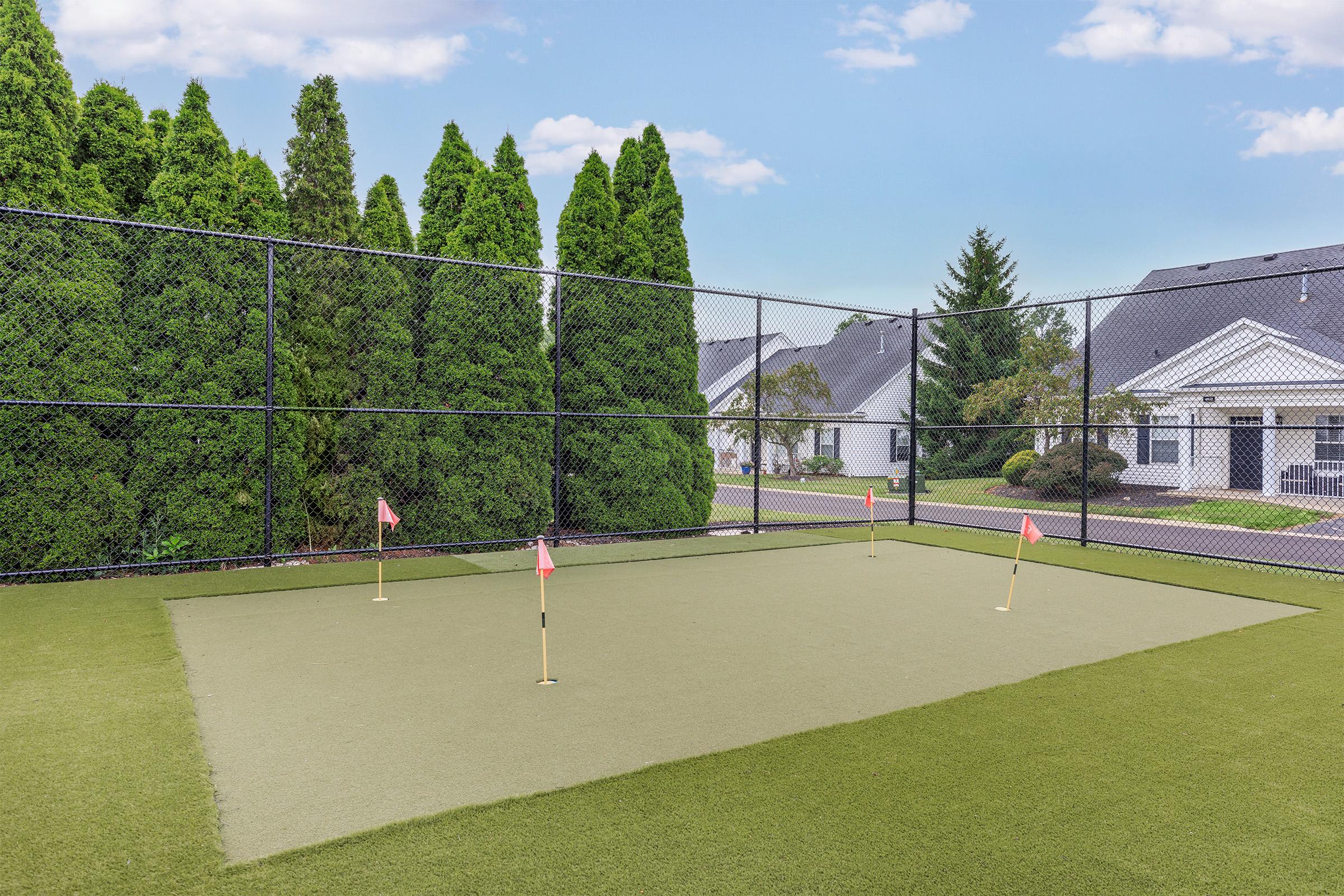 A well-maintained artificial turf practice putting green enclosed by a black fence, featuring several small flags marking the holes. Lush green trees are visible in the background, along with residential homes. The sky is clear with a few clouds, creating a serene outdoor environment.