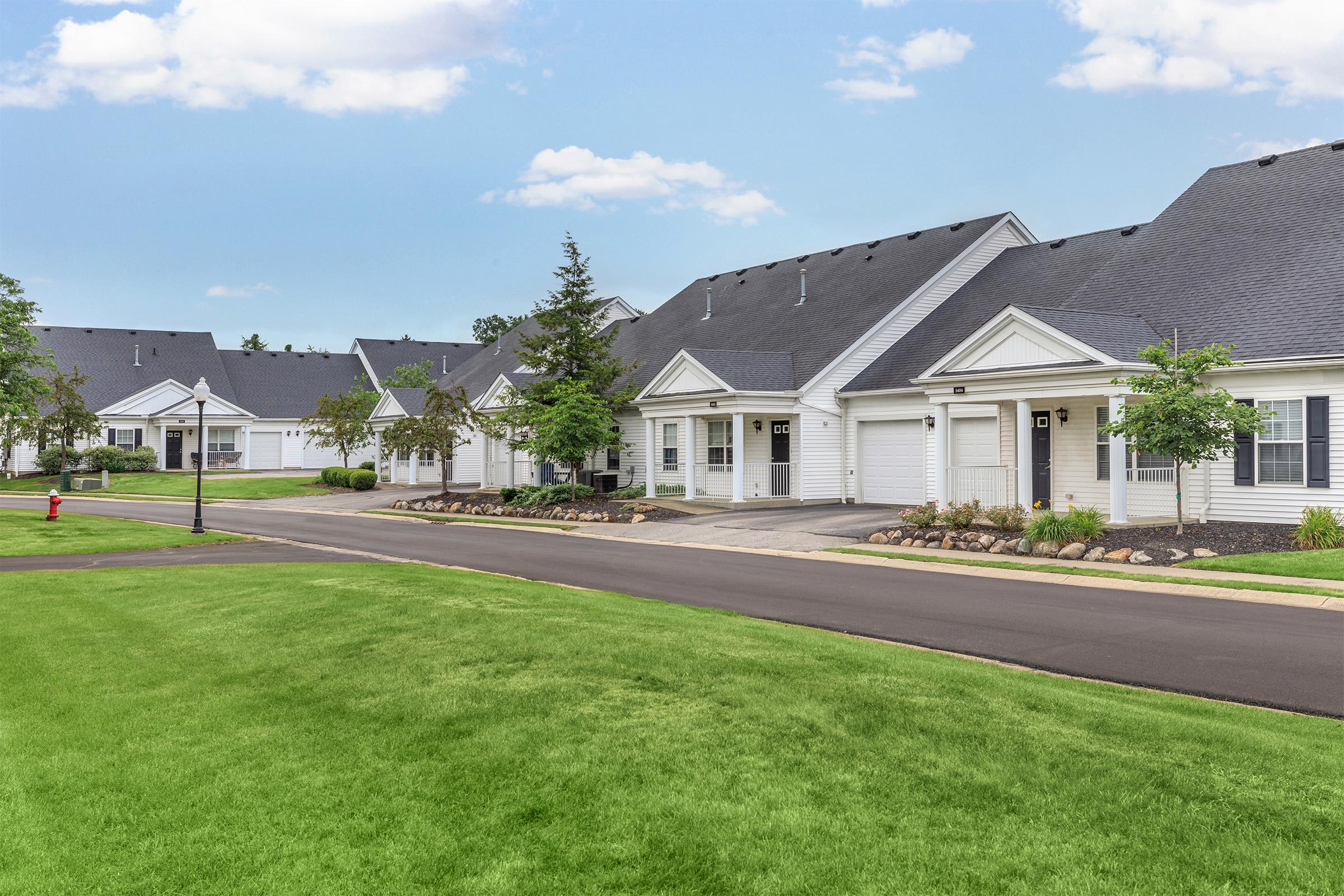 A row of well-maintained single-family homes with gray roofs and white siding, set in a landscaped area with green grass and small trees. The street is paved, and there are flower beds along the sidewalk. A fire hydrant is visible on the side of the road. The sky is clear with a few clouds.