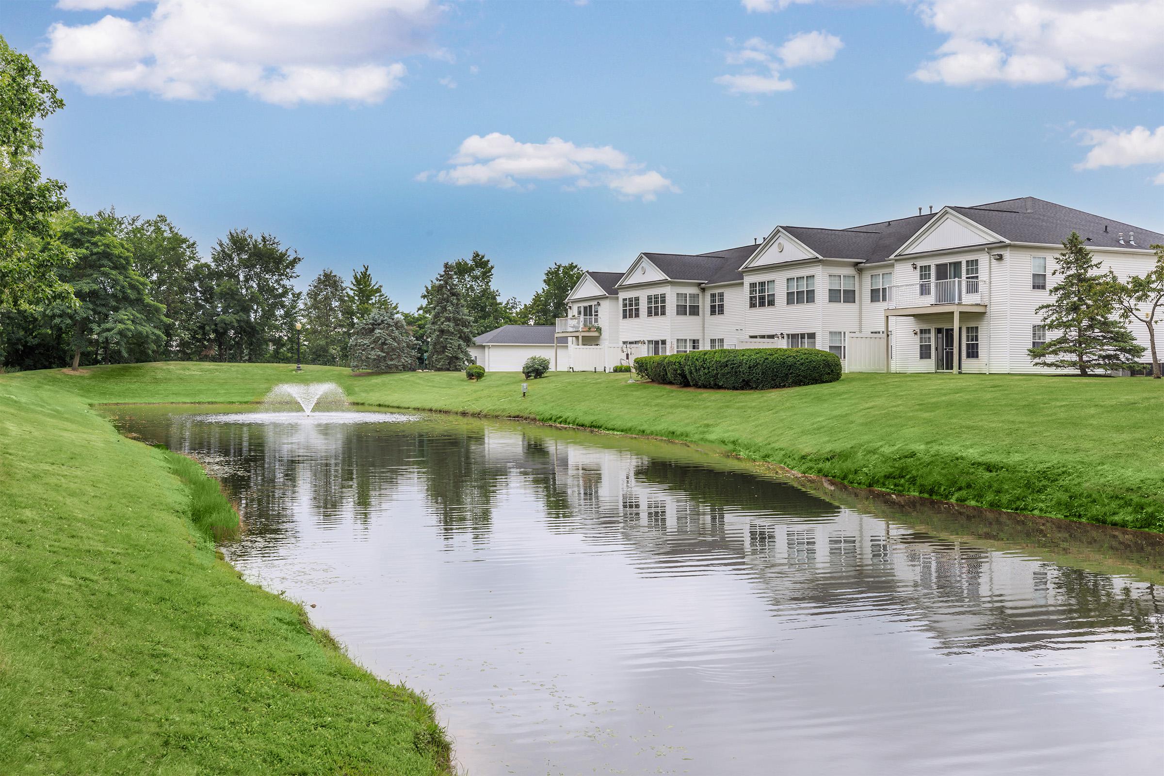 A serene landscape featuring a calm pond with a fountain, surrounded by lush green grass. In the background, several residential buildings with white facades and dark roofs are visible, framed by trees under a partly cloudy blue sky. The scene conveys a peaceful residential setting.