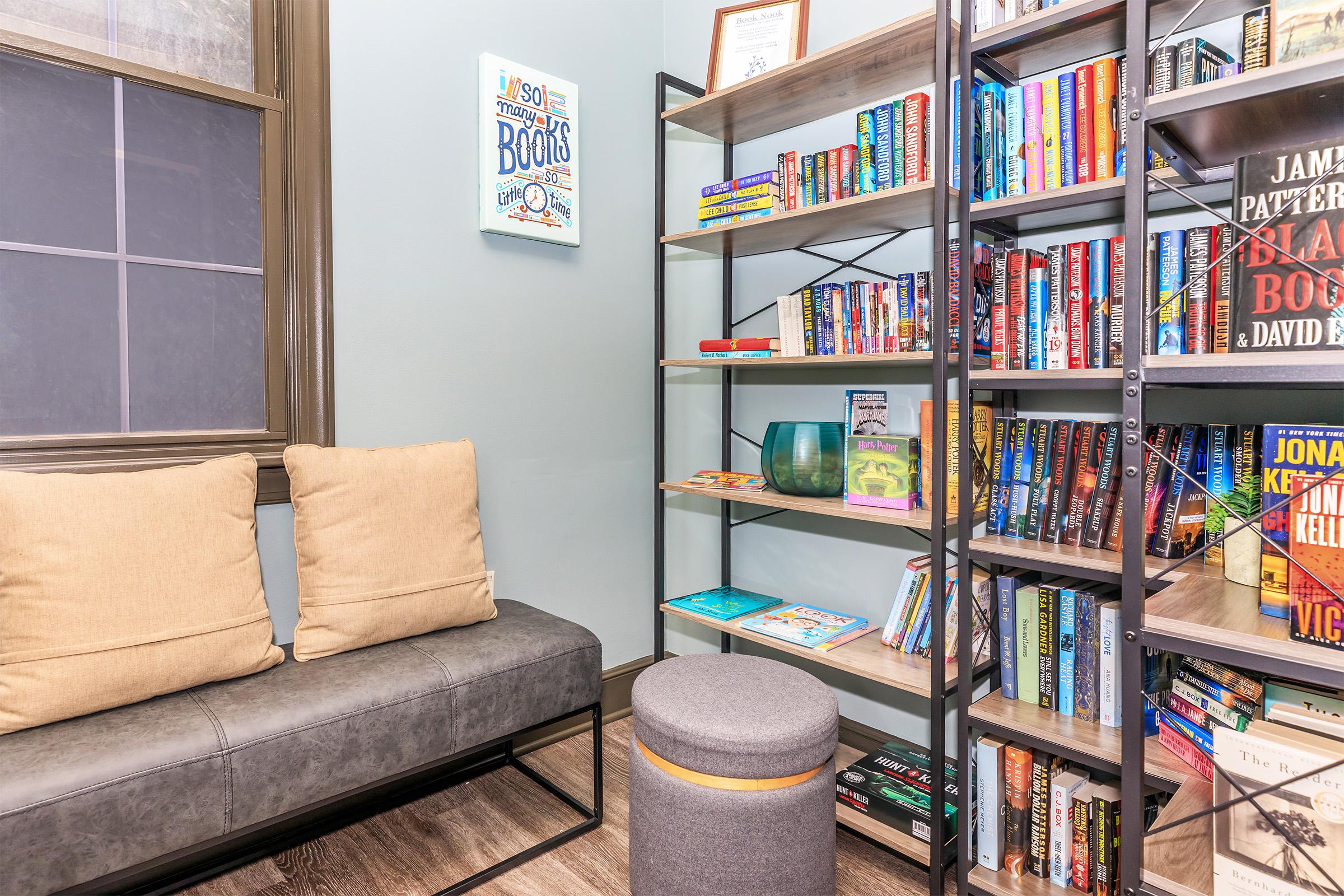 Cozy reading nook featuring a gray upholstered bench with two cushions, a round ottoman, and a modern shelving unit filled with colorful books. A framed poster about books hangs on the wall, and decorative items, including vases, are displayed on the shelves, all set against a light blue wall.