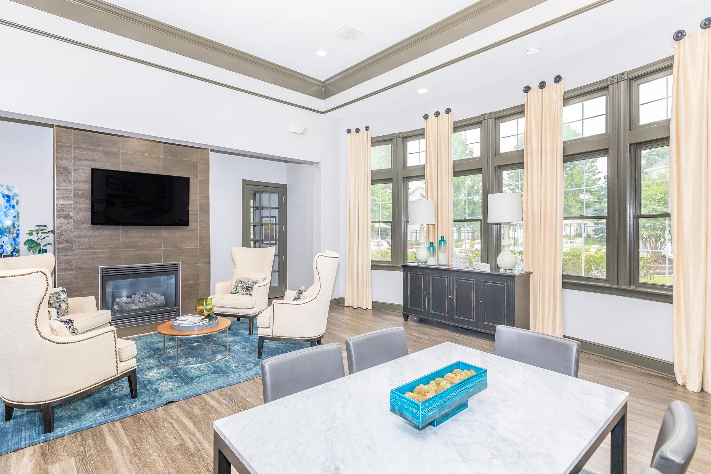 A modern living room featuring two elegant armchairs and a coffee table in front of a stone fireplace. Large windows with beige curtains allow natural light, and a dining table with a blue centerpiece is visible. The decor combines contemporary and cozy elements in a bright, welcoming atmosphere.