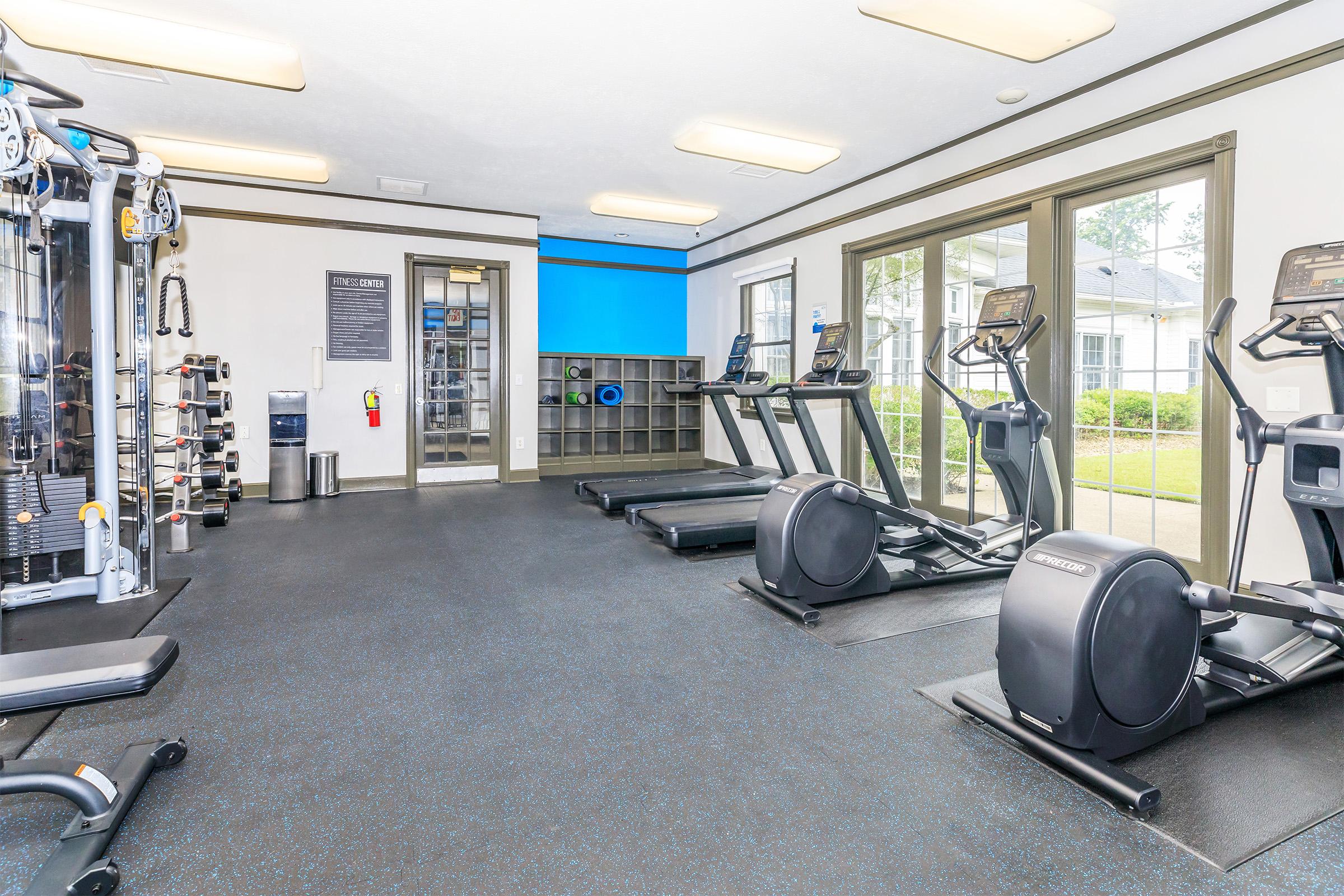 A modern gym interior featuring various fitness equipment, including treadmills, elliptical machines, and a cable machine. The space has rubber flooring, large windows for natural light, and a wall-mounted information board. Shelving displays exercise accessories, creating an inviting workout environment.