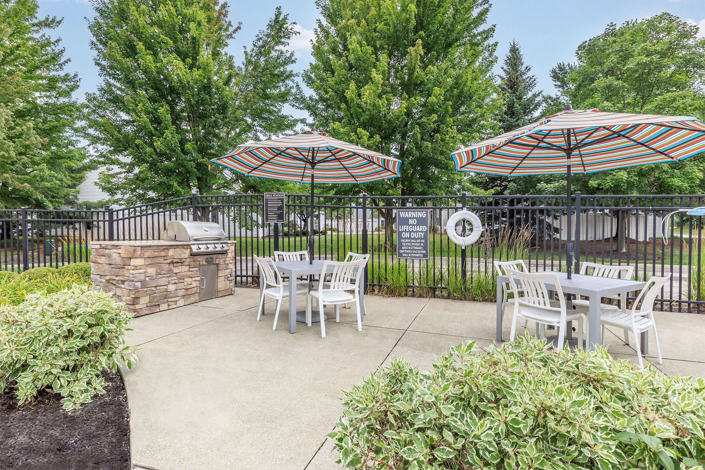 Outdoor patio area featuring a grill, white tables and chairs, and colorful striped umbrellas. Surrounding greenery and a safety sign are visible, enclosed by a black fence. The setting is designed for casual dining and relaxation.