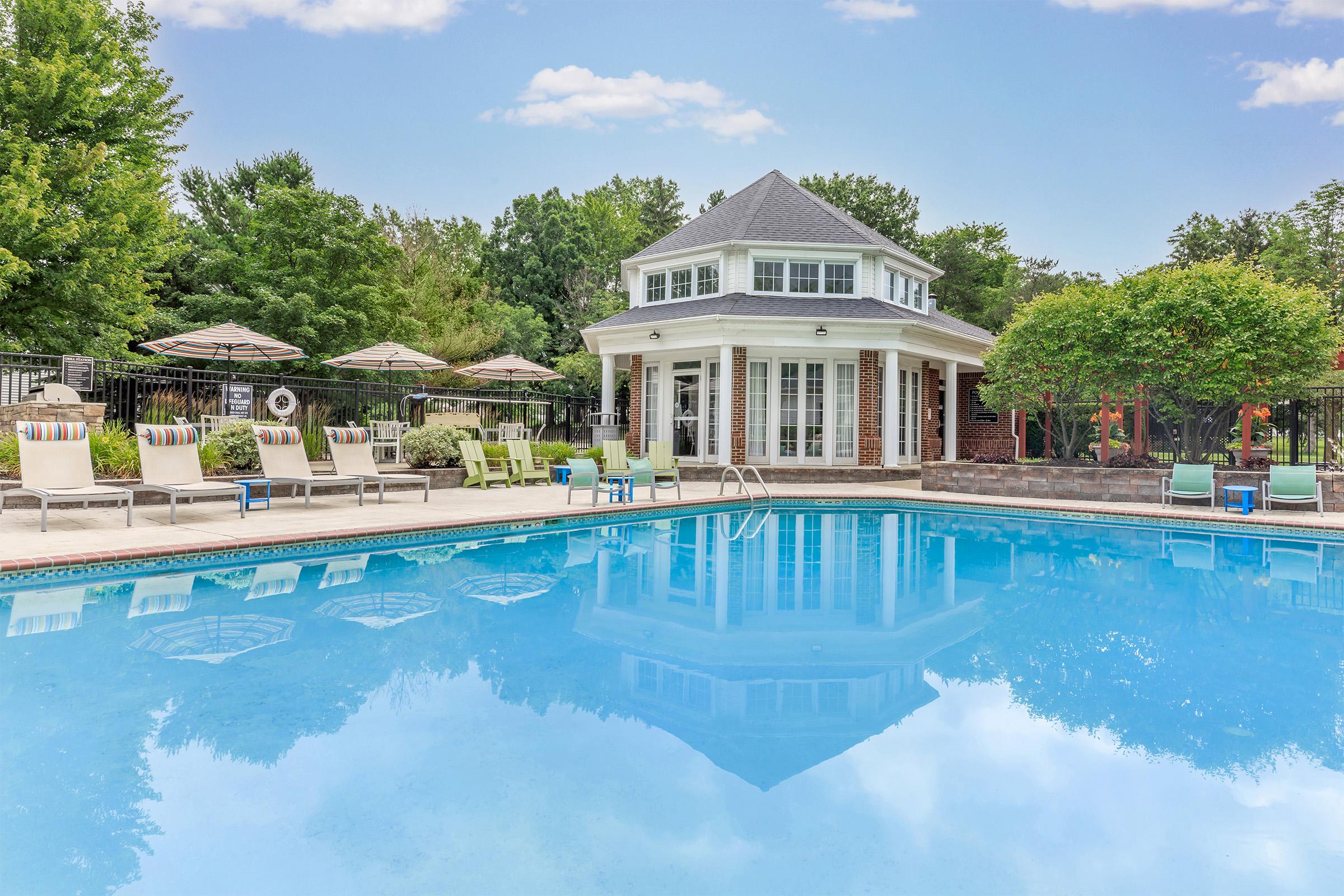 A serene swimming pool surrounded by lounge chairs and umbrellas, with a picturesque two-story building in the background. The water is crystal clear, reflecting the trees and sky, creating a tranquil atmosphere. Ideal for relaxation and leisure activities.