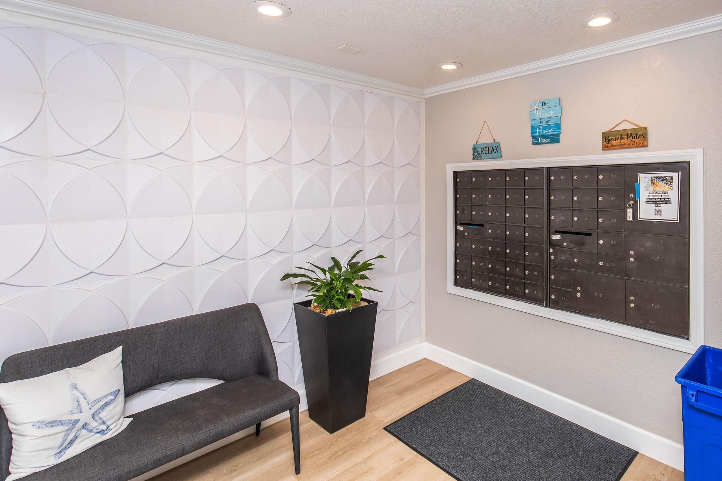 A modern lobby area featuring a light circular patterned wall, a dark mailbox unit mounted on the right with various mail slots, a decorative plant in a tall pot, a gray bench with a starfish pillow, and a blue recycling bin on the floor. Natural light is provided by ceiling lights.