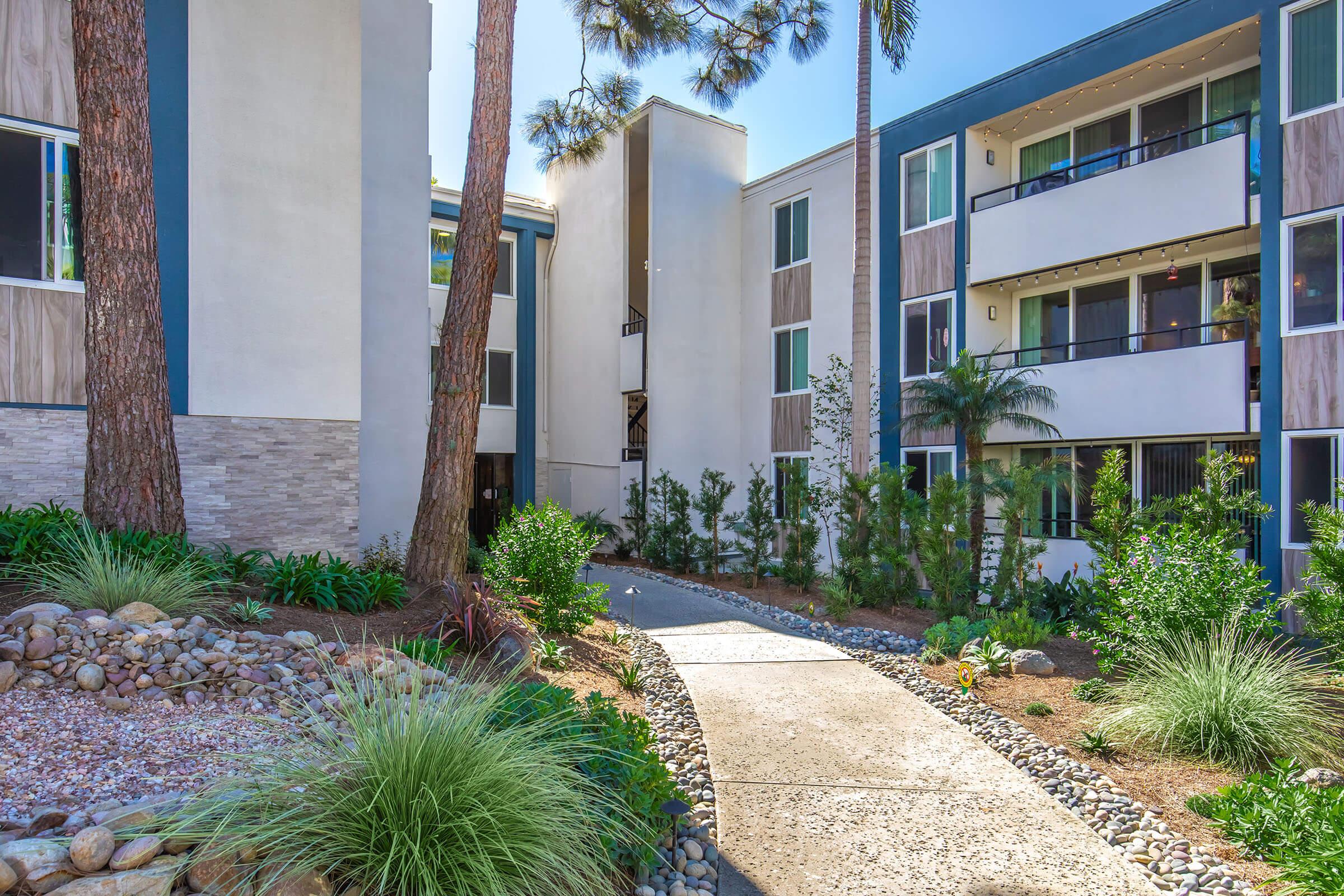 A landscaped pathway leads through a modern residential complex, framed by greenery and palm trees. The buildings feature a mix of white and wood paneling, with balconies visible. Sunlight illuminates the area, creating a warm and inviting atmosphere.