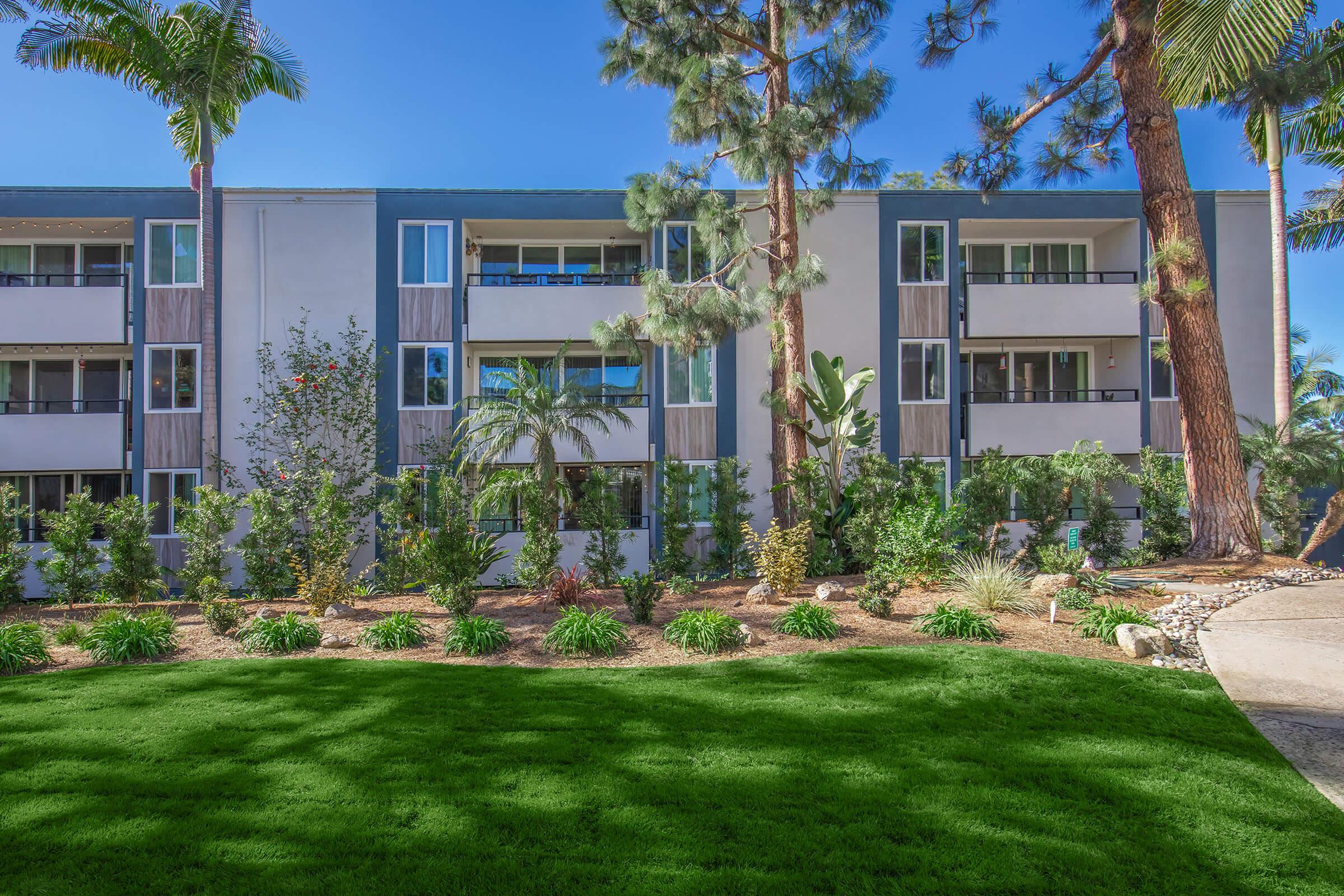 A modern apartment building surrounded by lush greenery and palm trees. The exterior features a mix of light and dark panels, with balconies visible on each floor. The well-maintained garden in front has ornamental plants and a vibrant lawn, enhancing the inviting atmosphere of the property.