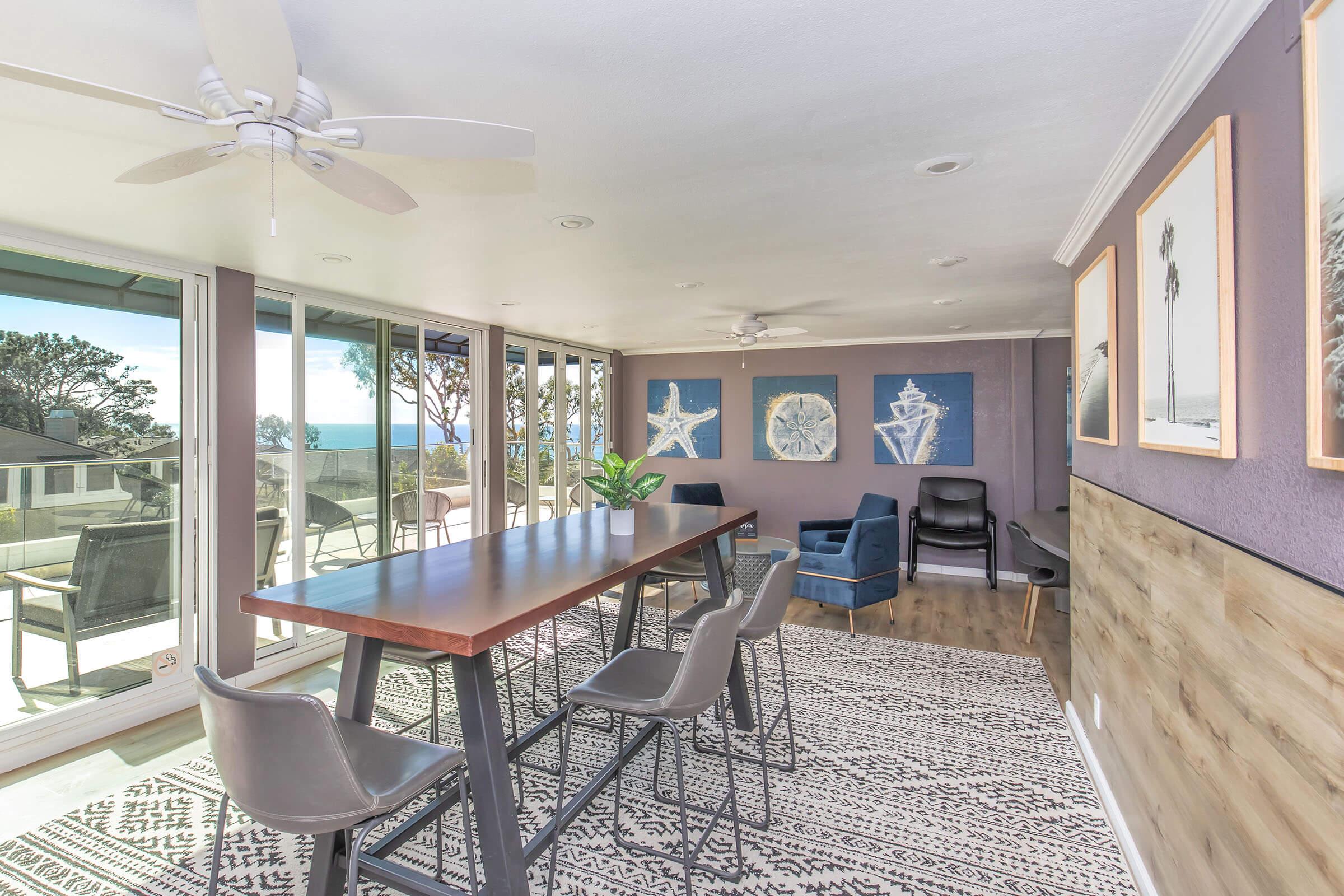 A spacious dining area featuring a long wooden table with chairs, large windows offering views of the ocean and surrounding landscape, and light-colored walls adorned with sea-themed artwork. A ceiling fan is visible, enhancing the airy atmosphere.