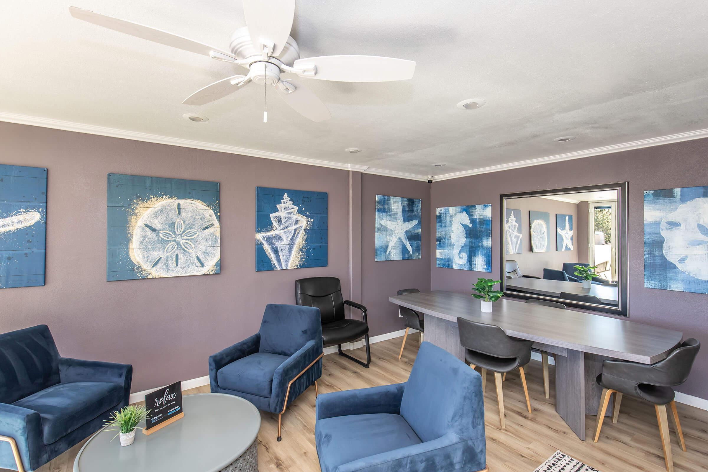 A modern, stylish room featuring a light purple wall, three blue chairs, a round coffee table, and a long dining table with black chairs. Blue artwork depicting various designs hangs on the walls, and a large mirror reflects the space. A ceiling fan adds to the contemporary atmosphere.