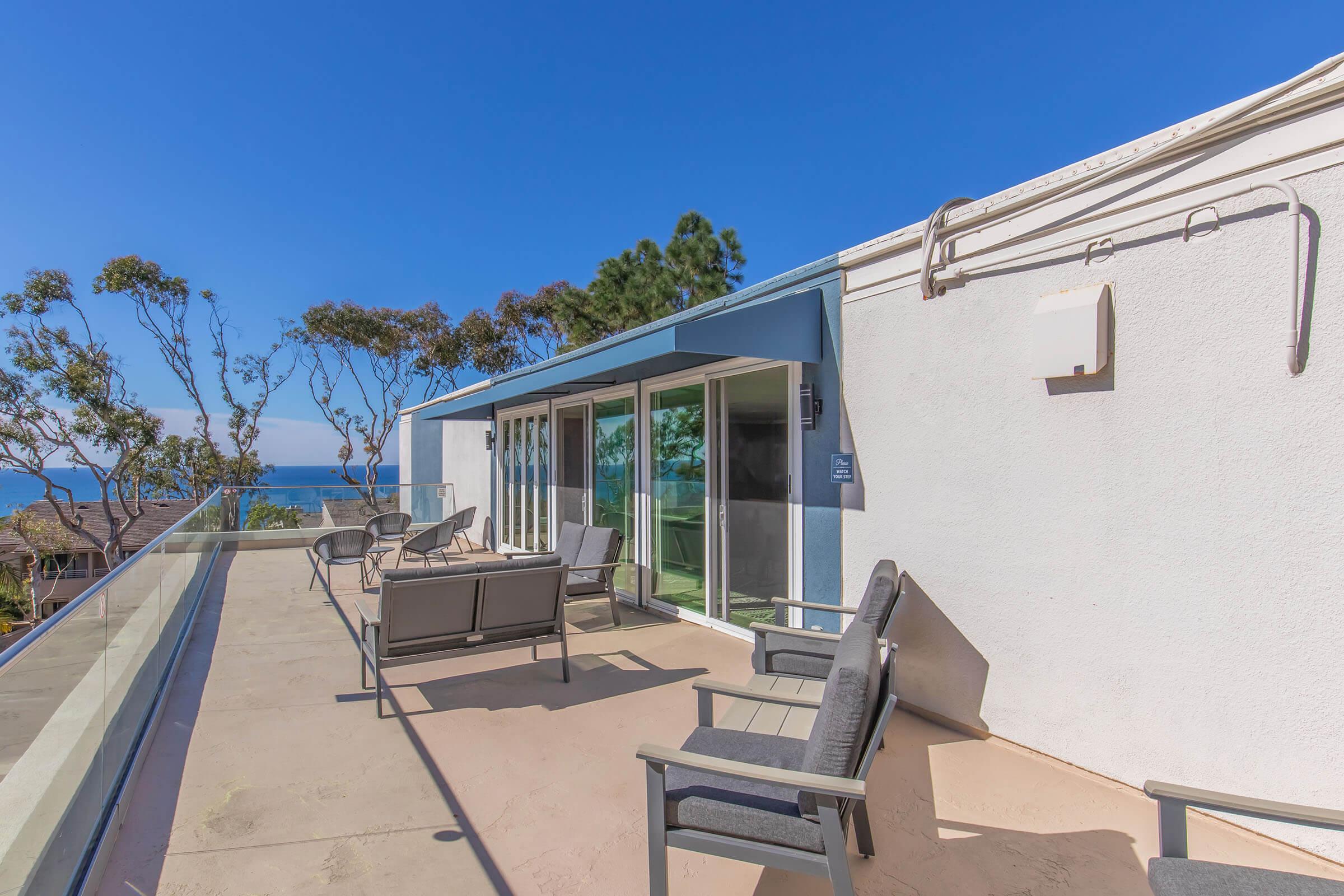 A sunlit patio overlooking the ocean, featuring several gray lounge chairs and large glass sliding doors. Surrounding greenery and trees add to the scenic view, with a clear blue sky above. The modern design creates a relaxed outdoor space perfect for enjoying the seaside atmosphere.