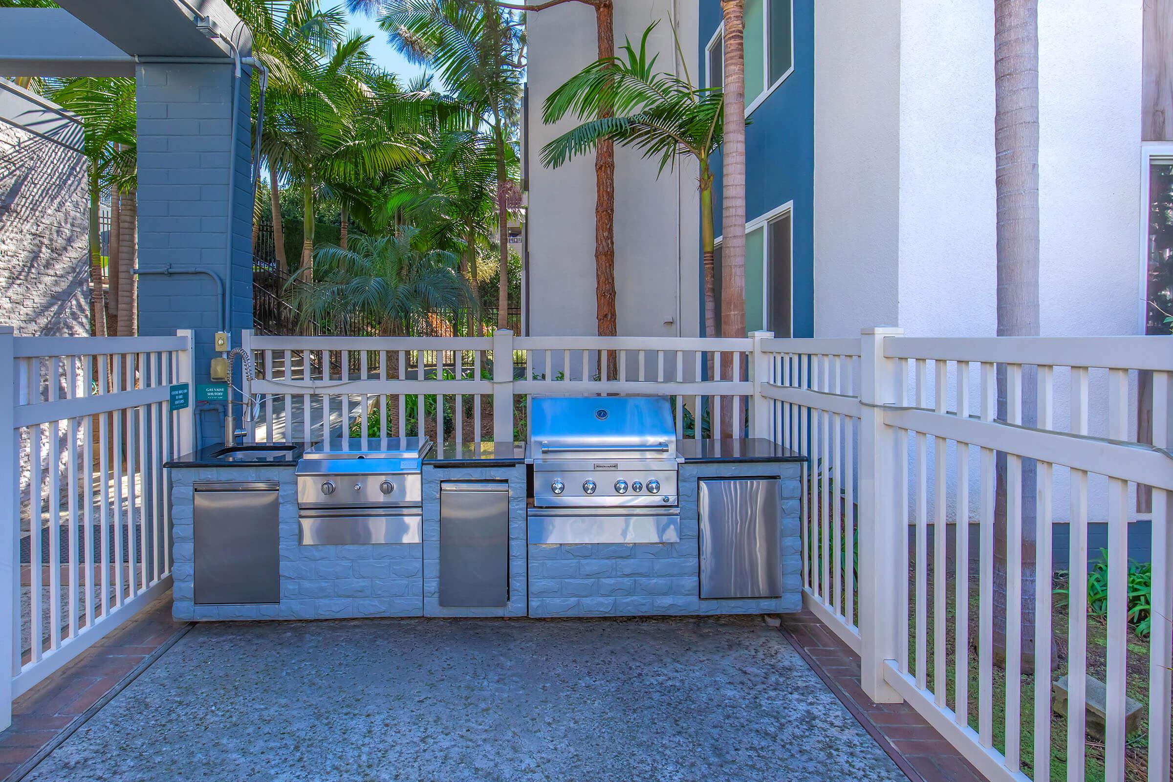 An outdoor grilling area enclosed by a white fence, featuring multiple stainless steel grills and countertops. The space is surrounded by lush green palm trees and modern buildings, creating a tropical ambiance for barbecuing.