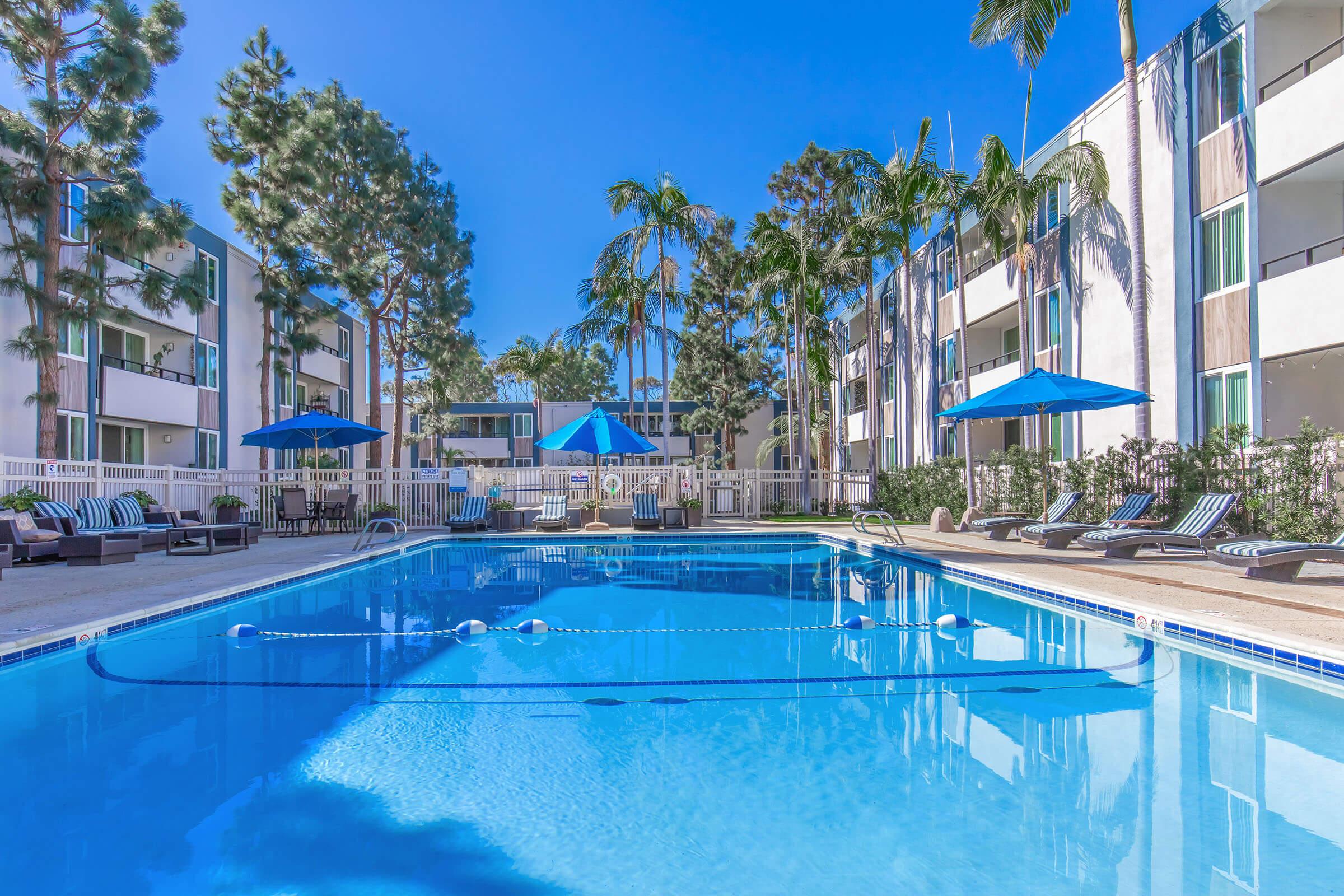 A serene swimming pool surrounded by palm trees and lounge chairs. Bright blue umbrellas provide shade. In the background, modern buildings with balconies can be seen under a clear blue sky, creating a tranquil and inviting atmosphere.