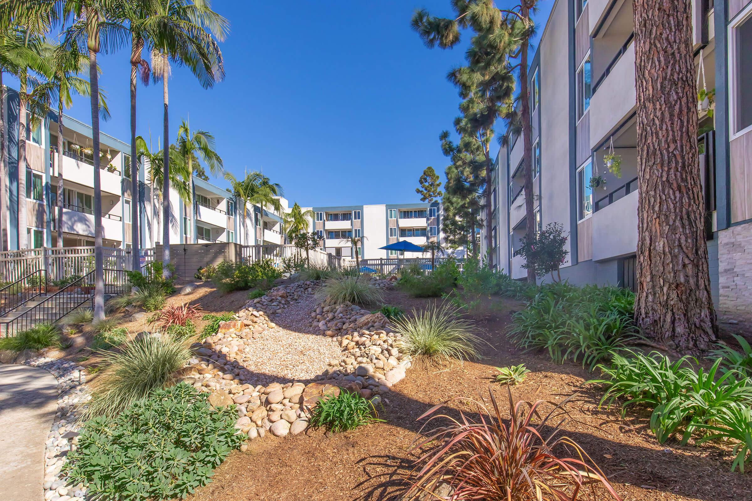 A landscaped pathway lined with greenery and rocks, leading to a courtyard with palm trees and an umbrella. Surrounding buildings feature multiple windows and a clear blue sky overhead. The overall scene conveys a serene and inviting atmosphere.