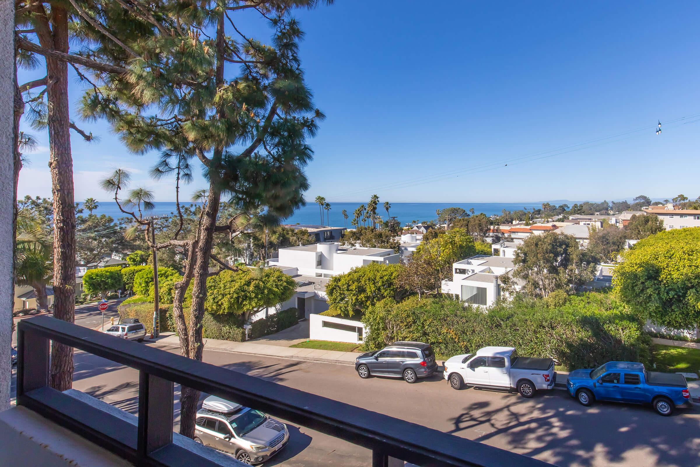 View from a balcony overlooking a coastal neighborhood. Lush green trees and well-maintained homes can be seen, with the ocean in the distance under a clear blue sky. Several parked cars line the street, adding to the tranquil suburban atmosphere.