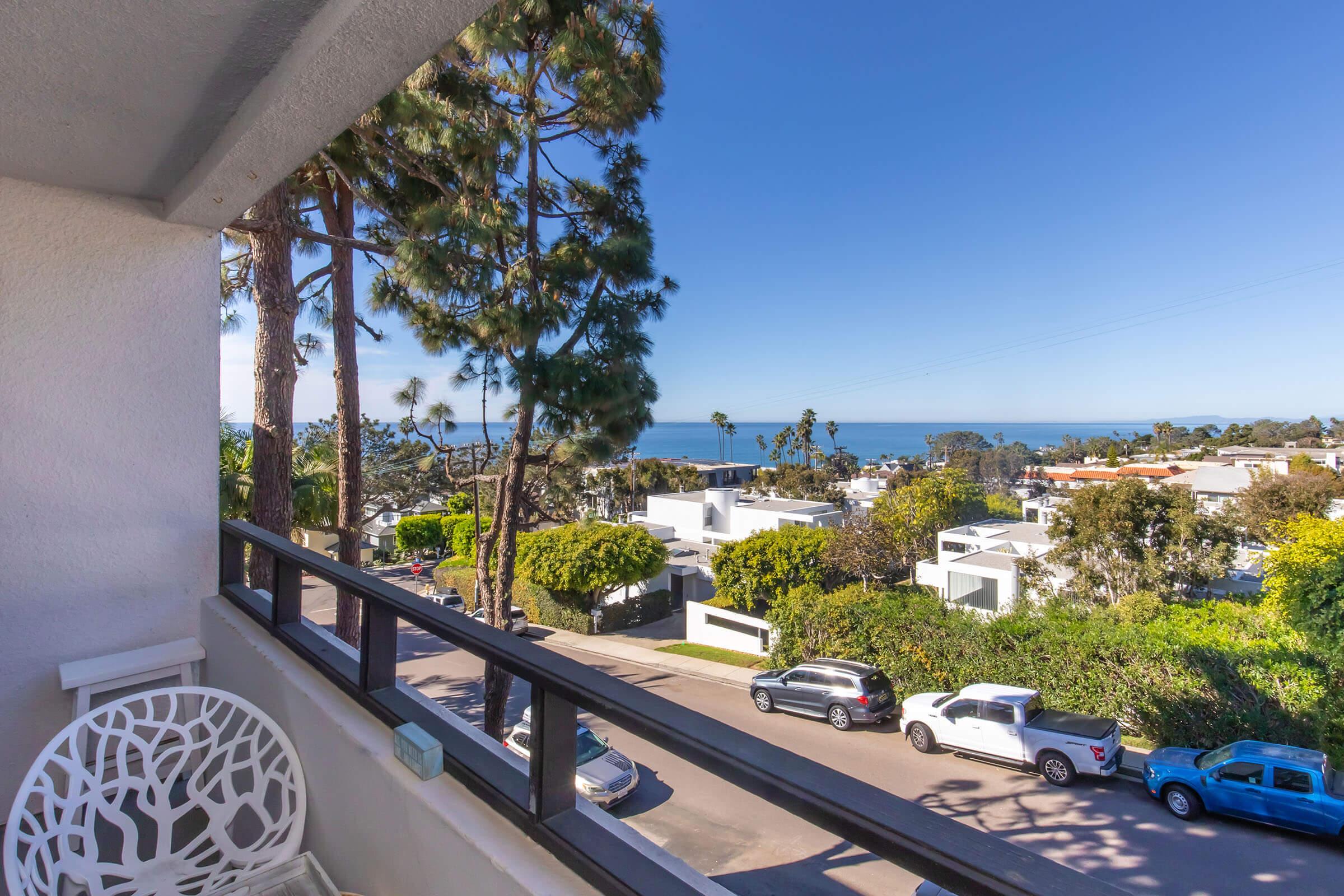 A scenic view from a balcony showcasing a coastal landscape. In the foreground, there are trees and modern houses, with a glimpse of the ocean in the background under a clear blue sky. Several vehicles are parked along the street, adding to the tranquil suburban atmosphere.