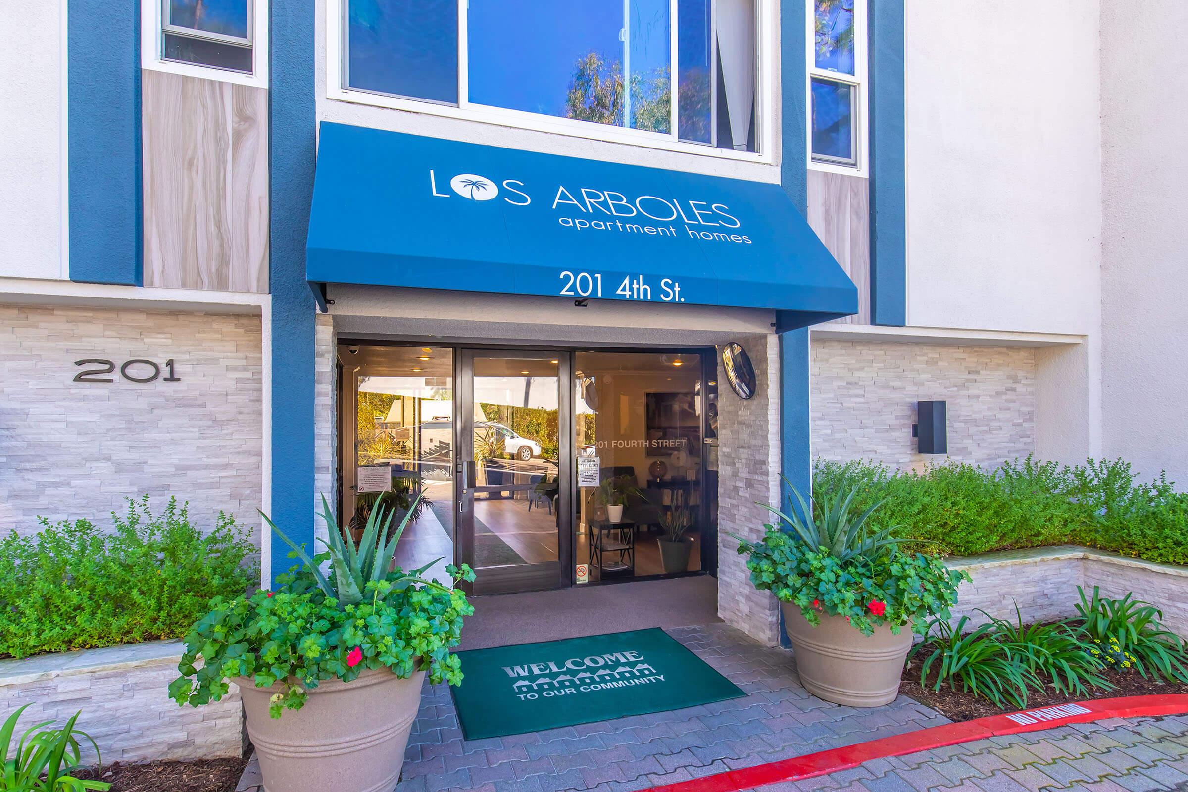 Entrance to Los Arboles Apartment Homes featuring a covered doorway with a sign indicating "201 4th St." and a welcome mat. It has decorative plants on either side and large windows that let in natural light, creating an inviting atmosphere.