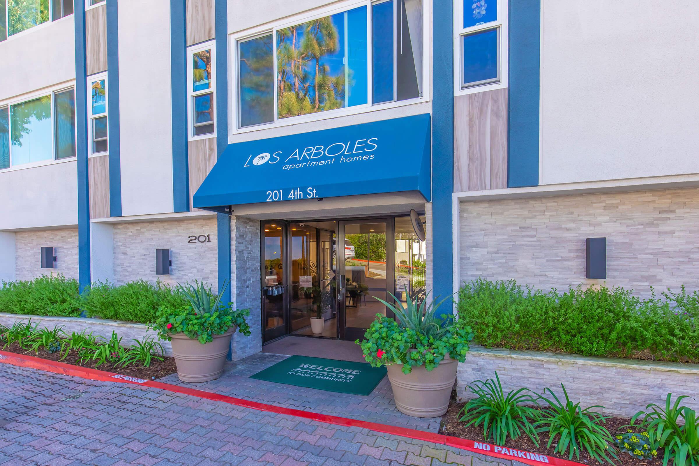 Entrance to Los Arboles Apartment Homes, featuring a blue awning with the name displayed. The building has large windows and modern architecture, surrounded by decorative plants and a welcoming mat. The address "201 4th St." is visible on the entrance.