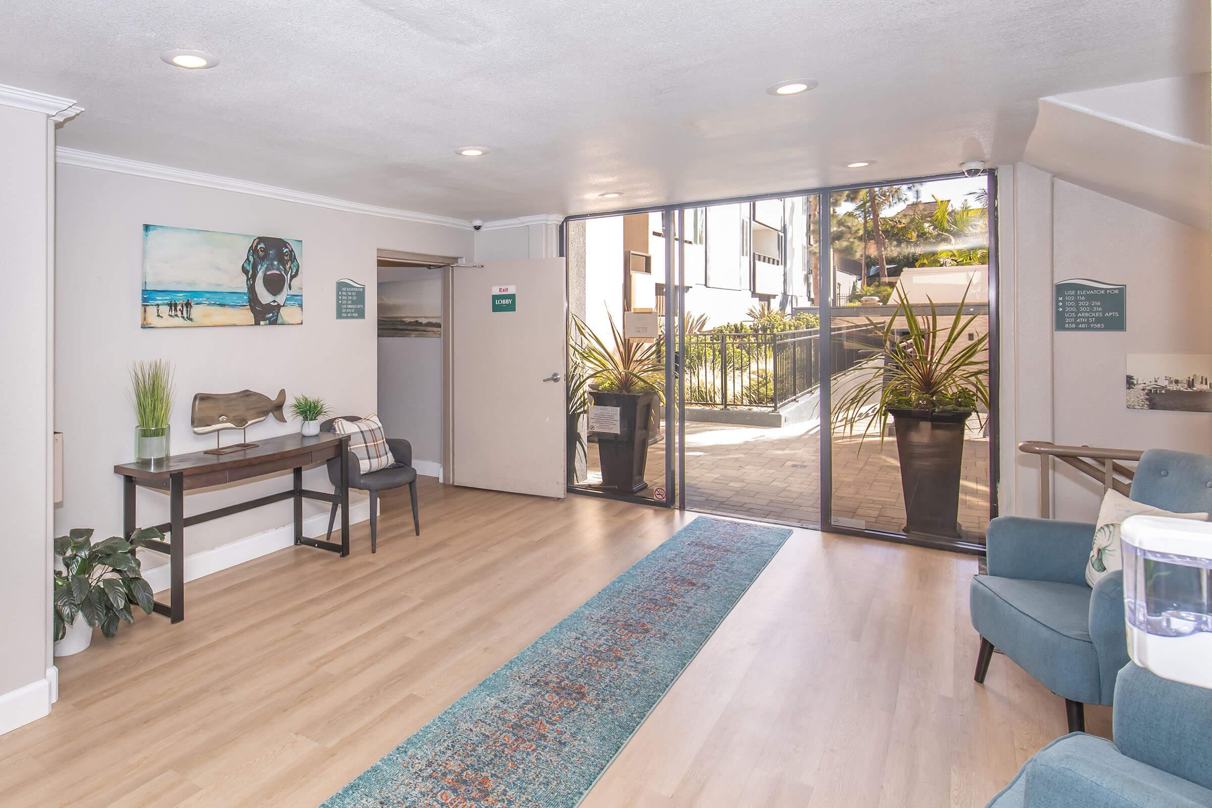 A bright and airy hallway featuring light wood flooring, contemporary furniture, and large windows revealing an outdoor area. Decor includes a painting of a dog, greenery in planters, and a small desk with a chair. Natural light enhances the inviting atmosphere of the space.