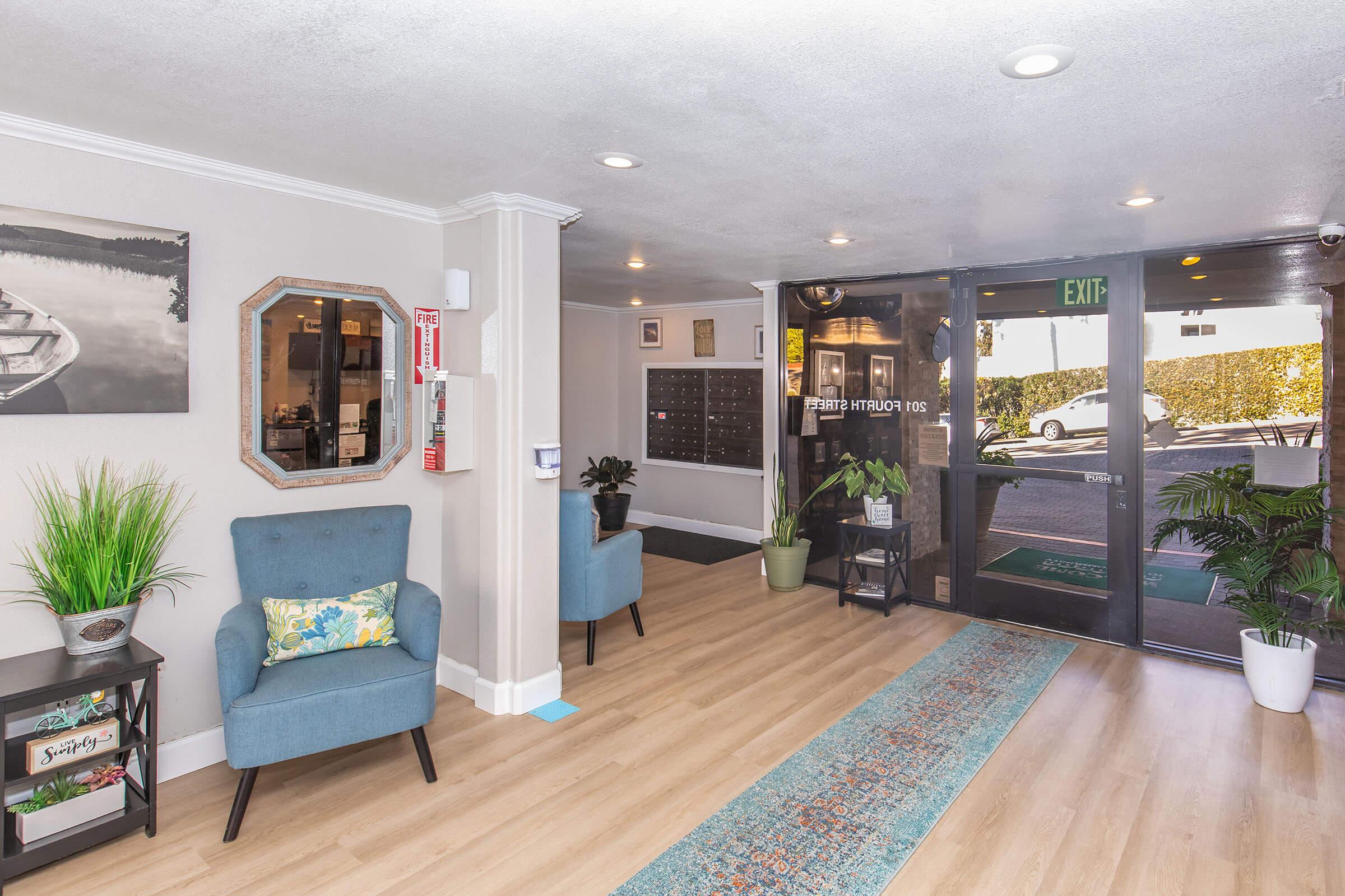 A welcoming lobby area featuring two blue chairs, a decorative mirror, a plant, and a colorful rug on light wood flooring. There is an entrance with glass doors leading outside, mailboxes on the wall, and a plant arrangement near the entrance. The space is well-lit and inviting.