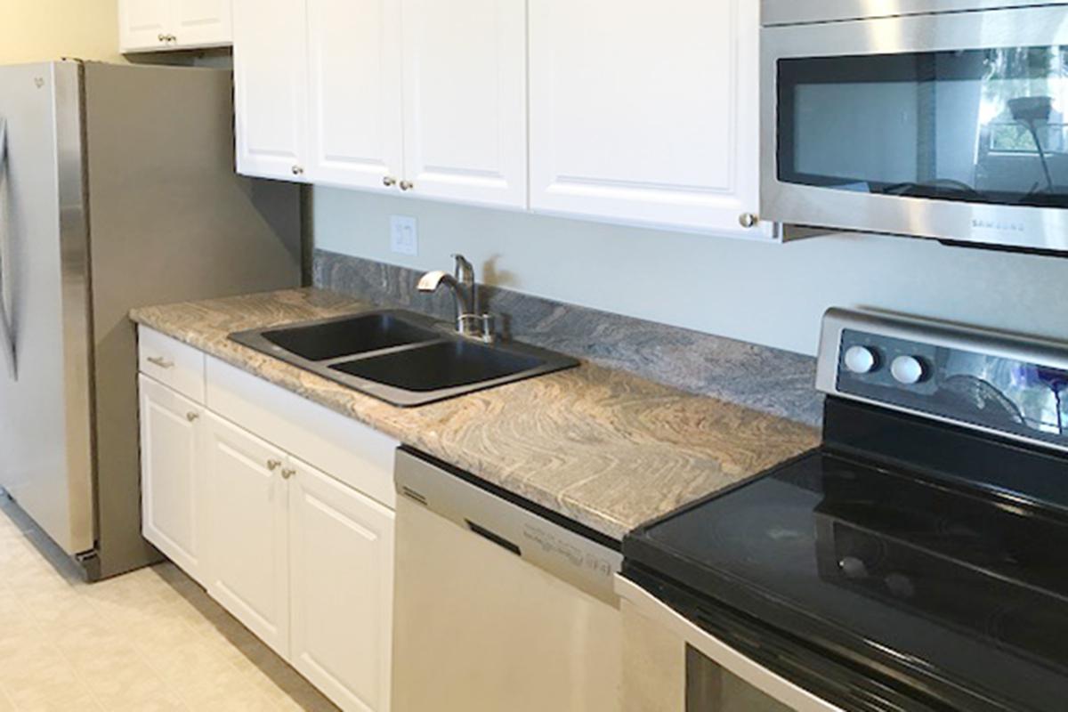 A modern kitchen featuring a stainless steel refrigerator, a microwave, a black oven, and a dishwasher. The countertop is made of granite with brown and gray patterns. There are white cabinets above and below the countertop, and a double sink with a sleek faucet. The flooring is light-colored tile.