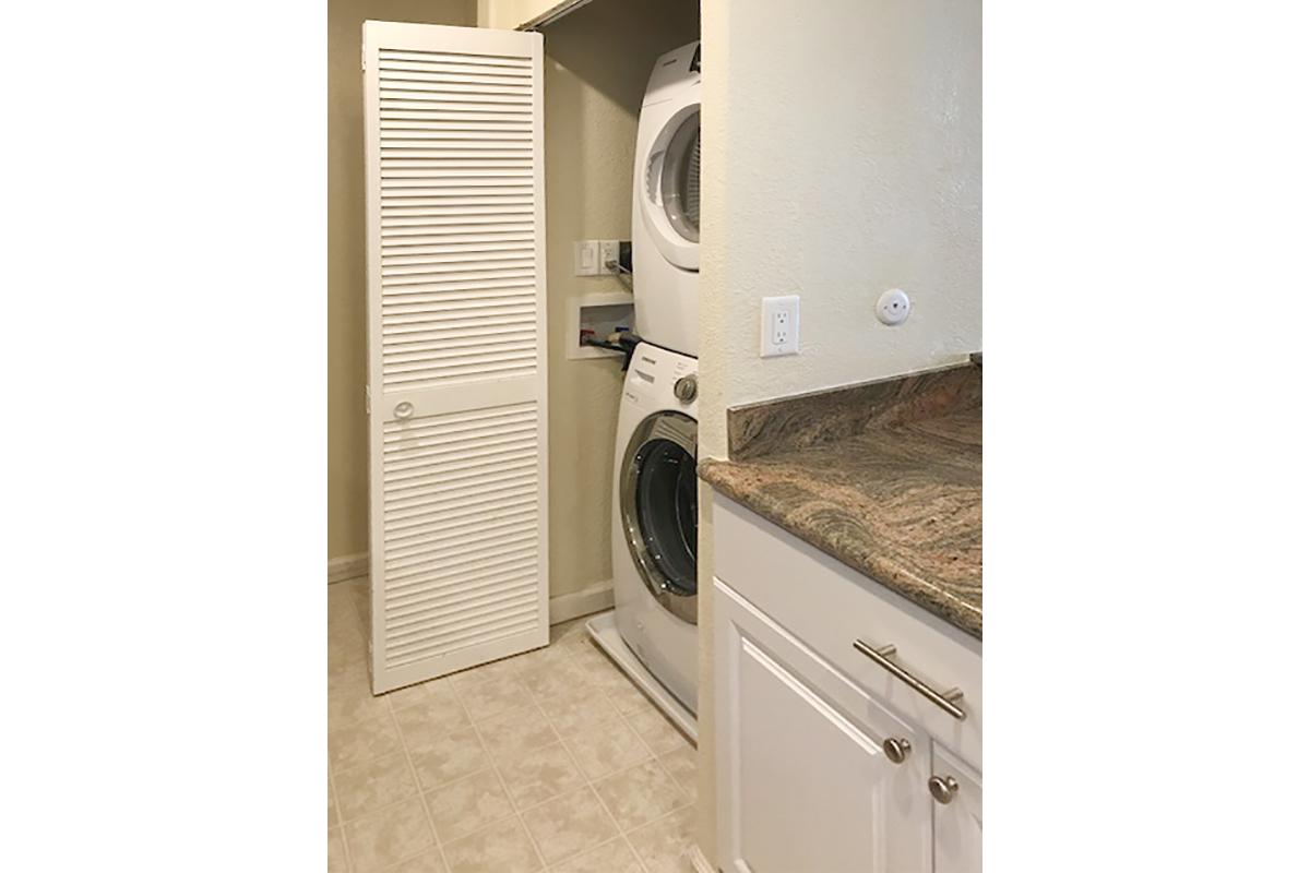 A small laundry area featuring a stacked washer and dryer in a closet, with light-colored walls and a granite countertop. There is a white cabinet door and beige tiled flooring, creating a clean and organized space.