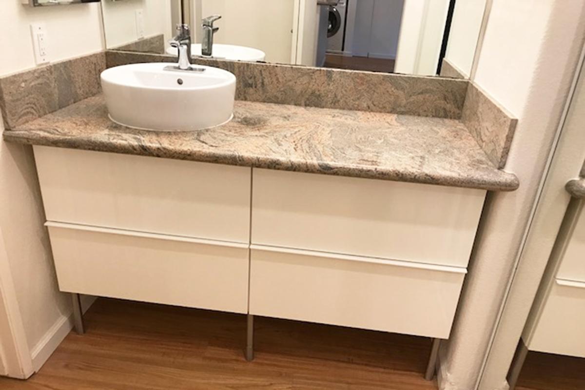 A modern bathroom vanity featuring a sleek white base with two drawers, topped with a large, stylish granite countertop. A round, white sink is centered on the countertop, complemented by a contemporary faucet. The background includes a glimpse of a bathroom mirror and neutral-colored walls.