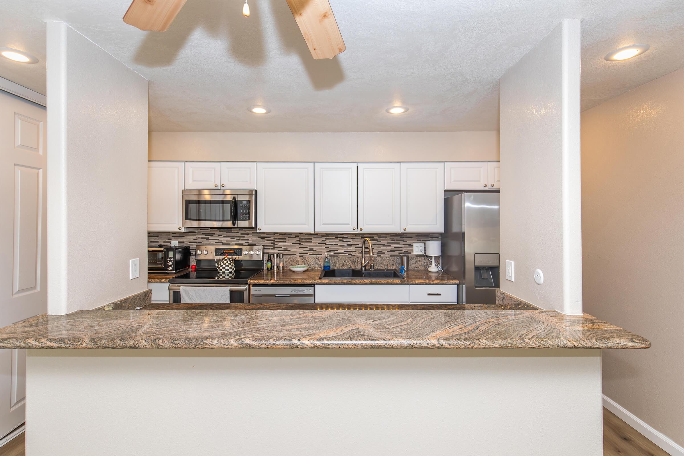 A modern kitchen with light-colored cabinetry, stainless steel appliances, and a decorative backsplash. The kitchen is partially open to a living space, featuring a granite countertop with seating. Soft lighting and a ceiling fan add to the inviting atmosphere.