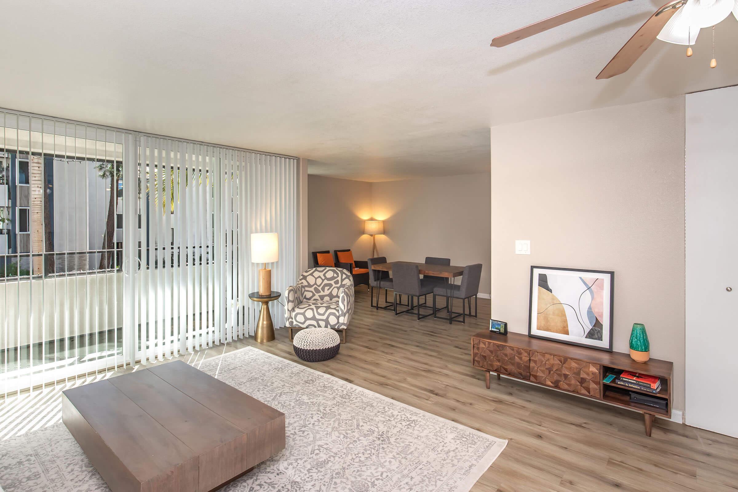 A stylishly furnished living room featuring a patterned armchair, a wooden coffee table, and a modern TV stand. Natural light streams through large windows with vertical blinds. In the background, there's a dining area with a table and chairs, creating an inviting atmosphere.