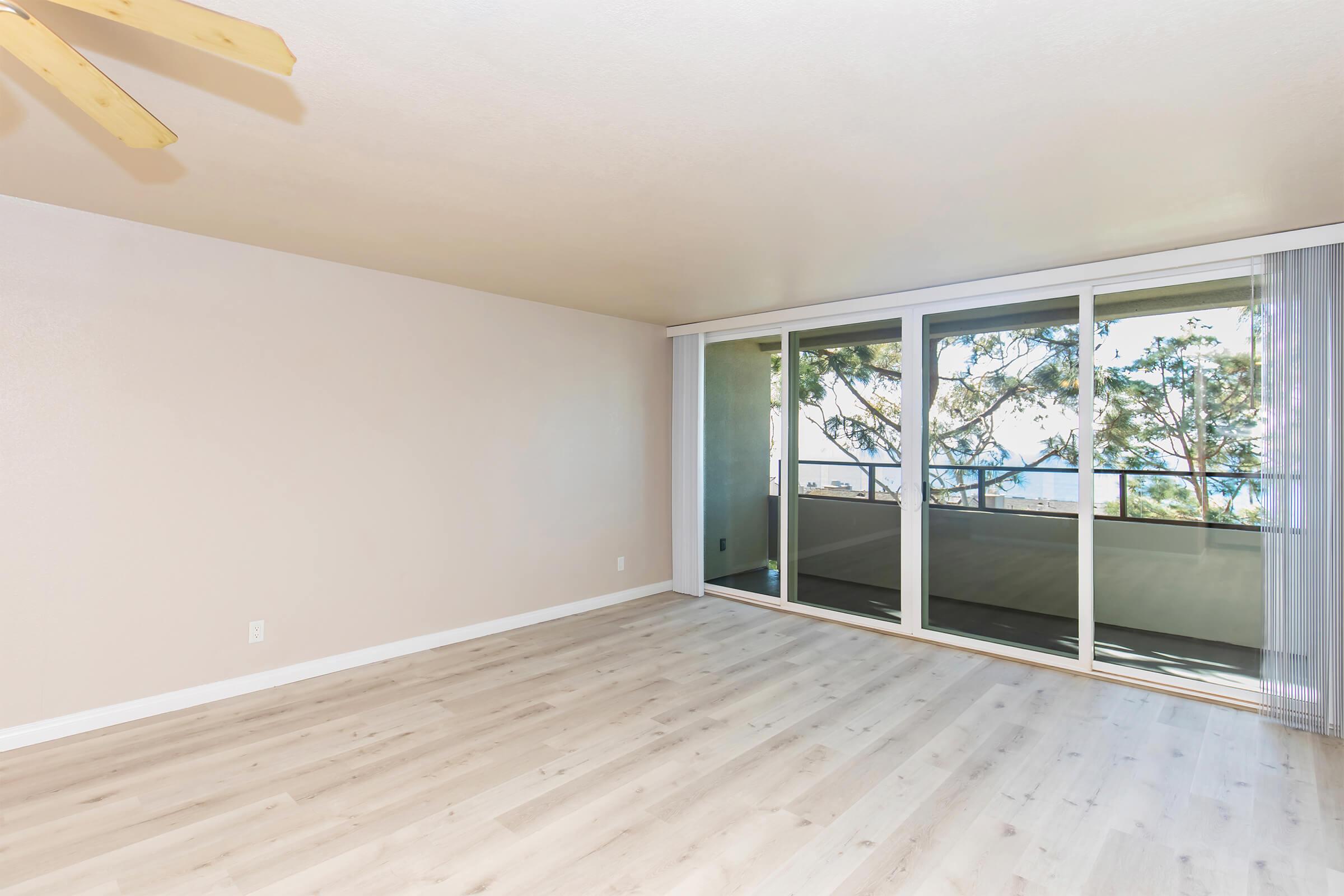 A spacious, empty room with light-colored walls and wooden flooring. A ceiling fan is visible, and large glass sliding doors open to a balcony with views of trees. Natural light fills the space, creating a bright and airy atmosphere.