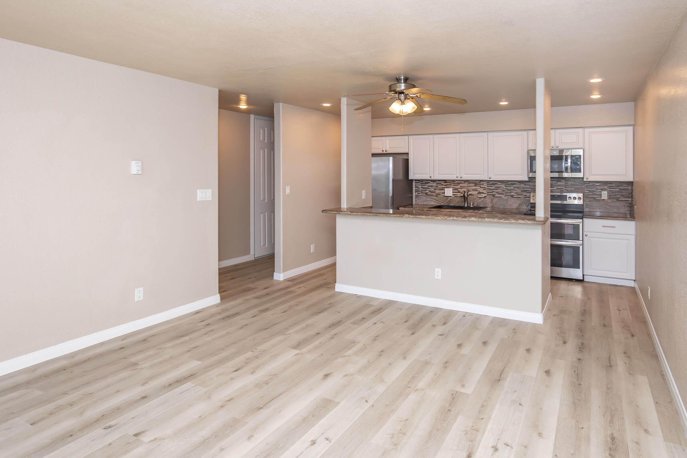 Spacious living area with light-colored wooden flooring, featuring a ceiling fan and a kitchen bar with modern cabinetry. The kitchen is partially visible, showcasing stainless steel appliances and a stylish backsplash. Neutral wall colors create a bright and open atmosphere.