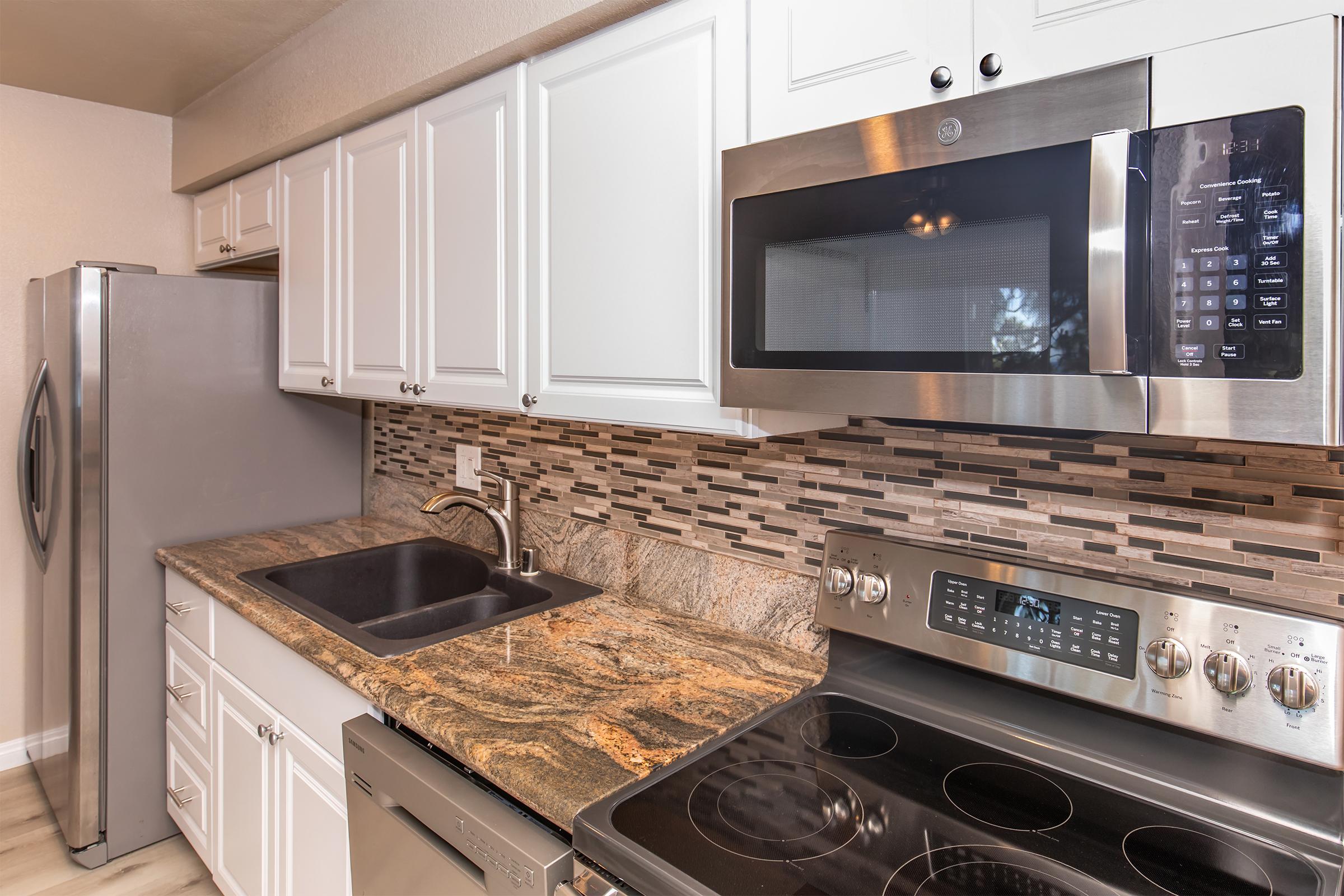 A modern kitchen featuring white cabinetry, stainless steel appliances including a refrigerator, microwave, and oven. The countertop is made of granite with a dark sink, and the backsplash consists of textured tiles in various shades. The overall design is bright and contemporary.