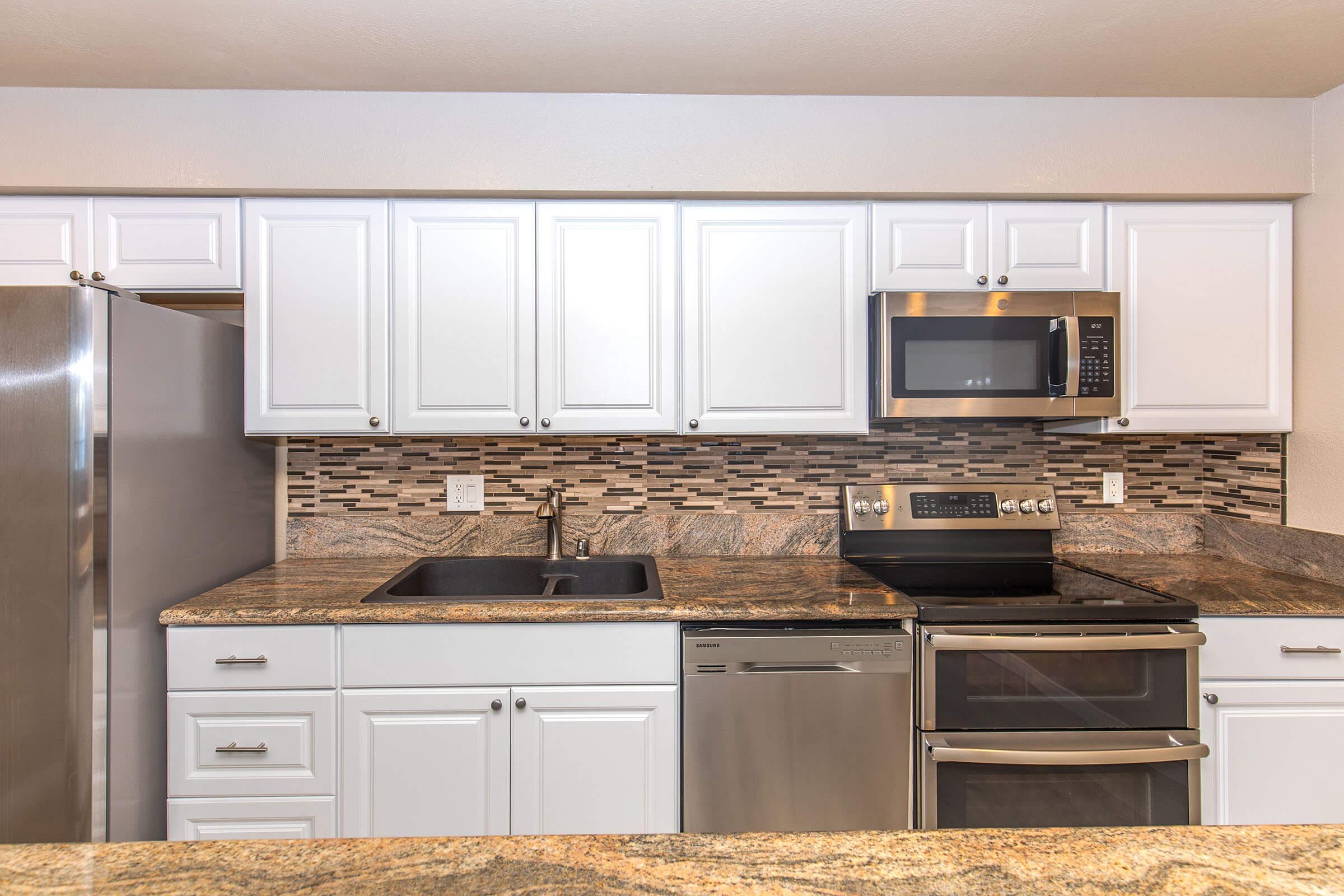 Modern kitchen featuring white cabinetry, granite countertops with a textured backsplash, stainless steel refrigerator, microwave, and oven. A double sink is installed, with a dishwasher underneath. The layout is open and bright, showcasing a contemporary design suitable for cooking and entertaining.