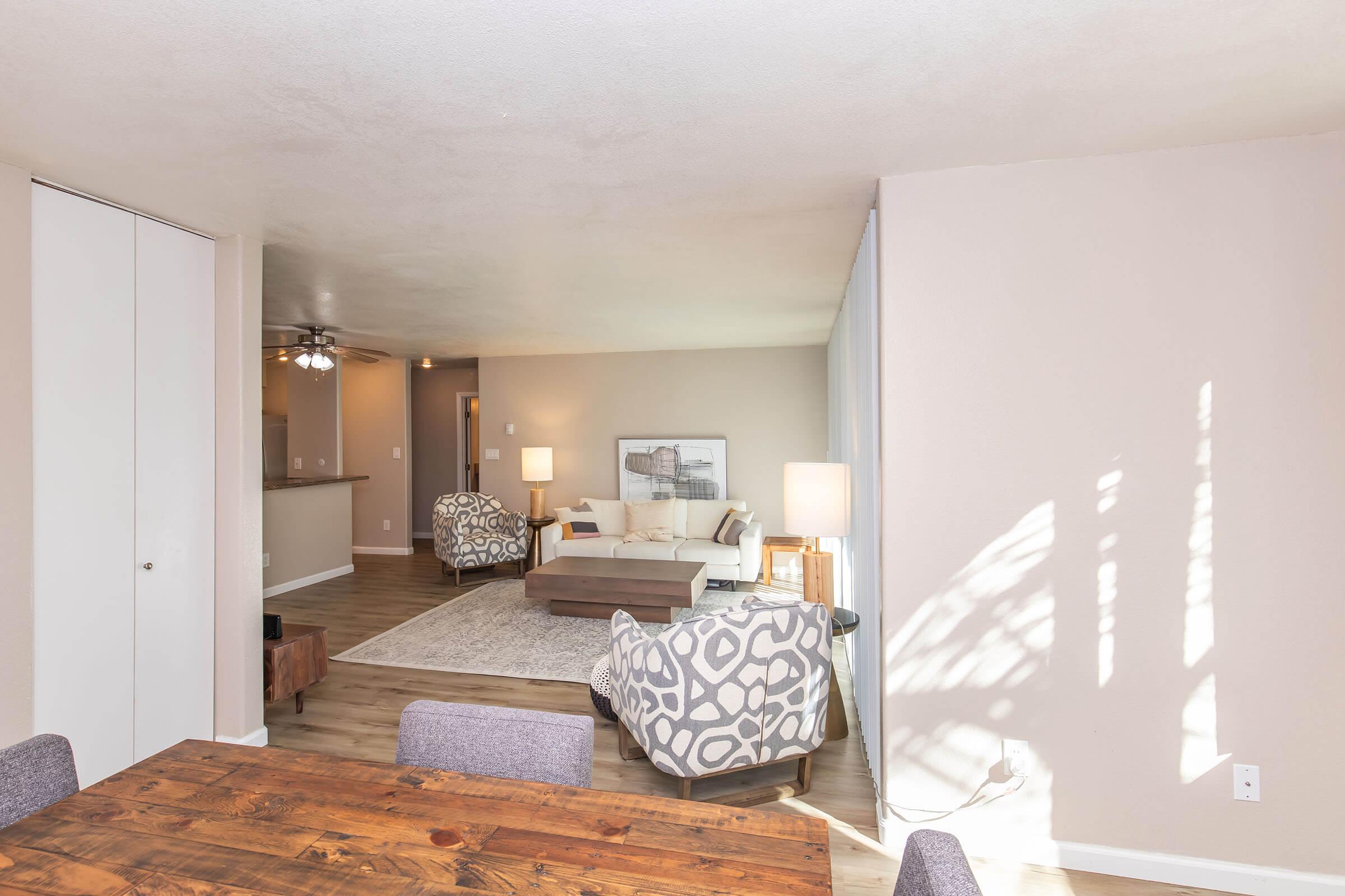 Spacious living area featuring a light-colored sofa and patterned chairs, with a wooden coffee table on a rug. The dining area includes a wooden table with chairs. Soft natural light fills the room, highlighting neutral walls and flooring. A wall-mounted lamp adds warmth to the ambiance.