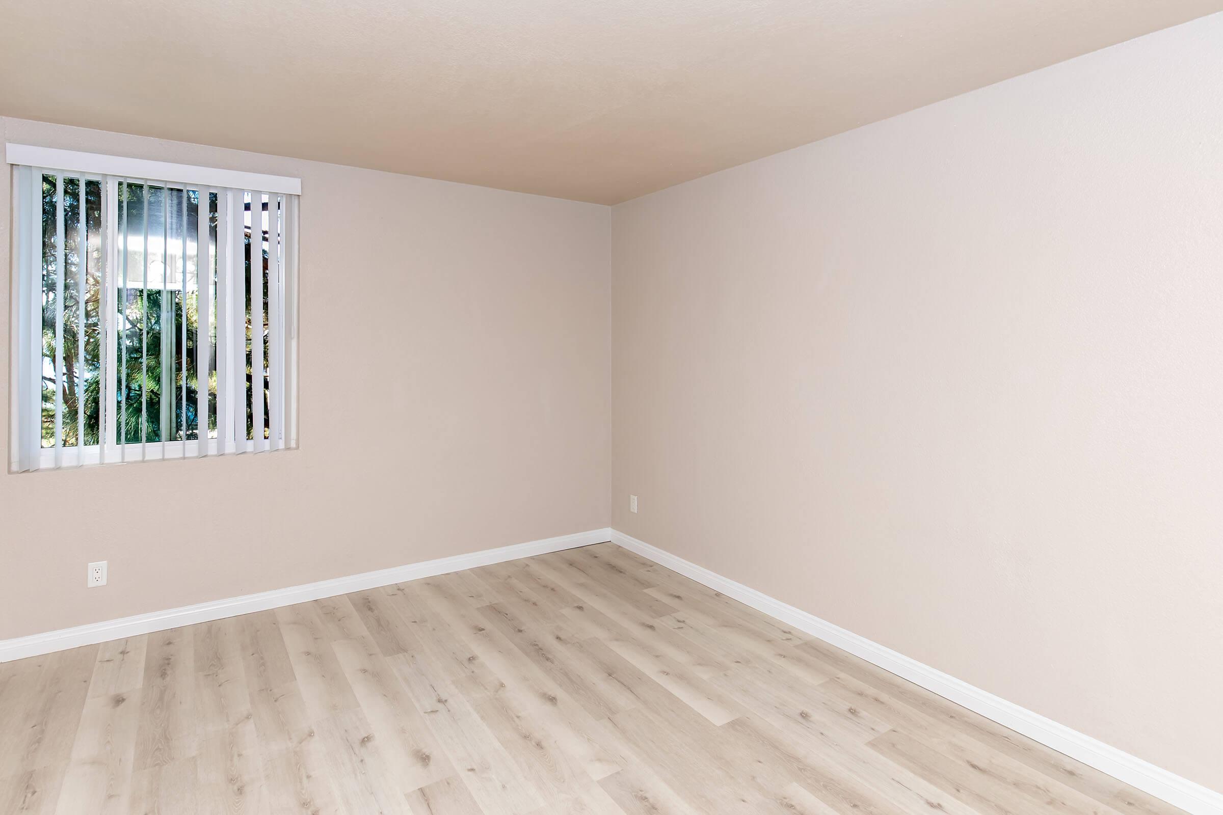 Empty room with light beige walls and laminate flooring. A window with vertical blinds allows natural light to enter. The walls are unadorned, creating a minimalist atmosphere.