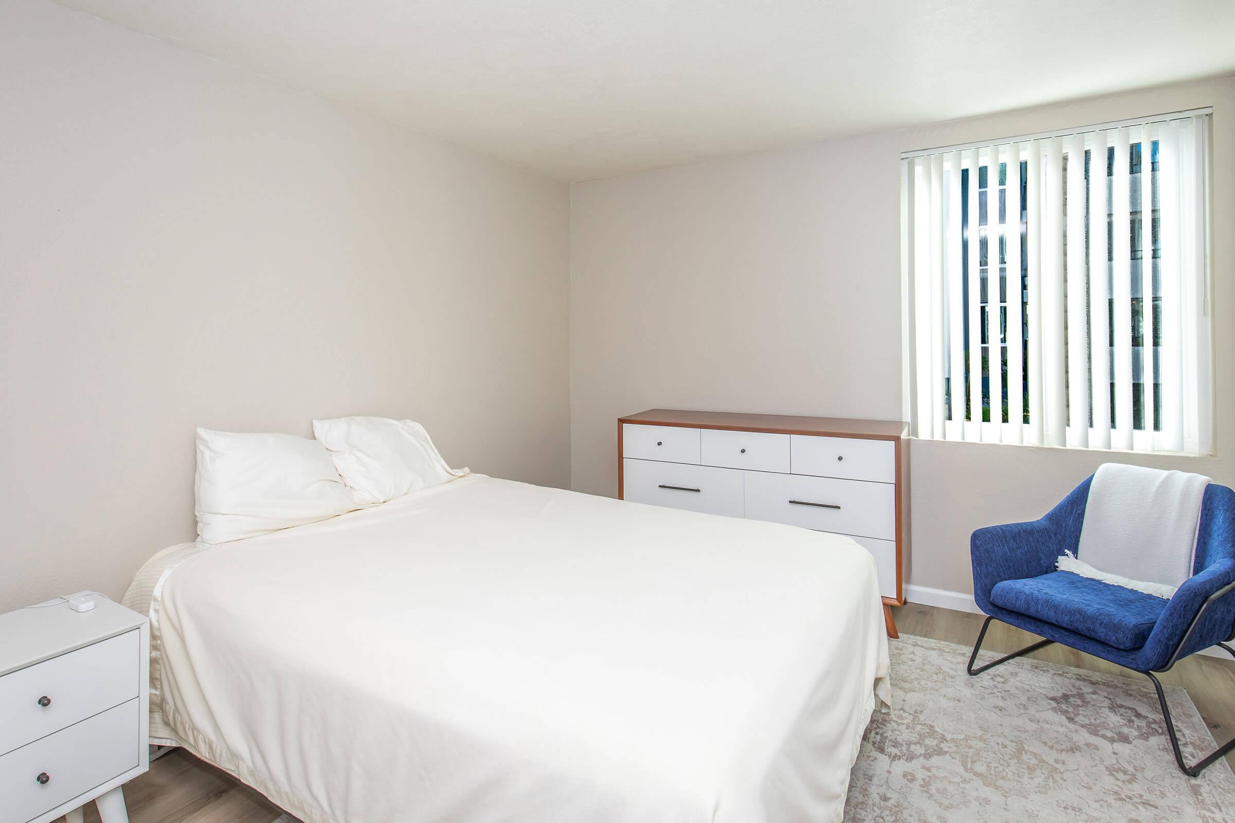 A minimalist bedroom featuring a white bed with a beige cover, a nightstand, a wooden dresser, and a blue accent chair. The walls are light-colored, and there are vertical blinds on a window, allowing natural light to enter the room. A soft area rug is placed under the chair.