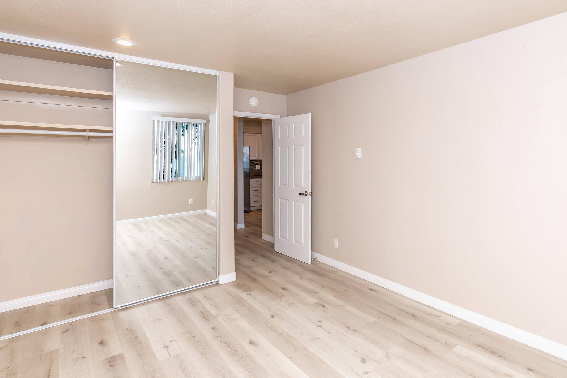A well-lit, minimalist room with light-colored walls and wooden flooring. There is a sliding mirror closet on one side and a white door leading to another space. Natural light comes from a window with vertical blinds, creating a spacious and inviting atmosphere.