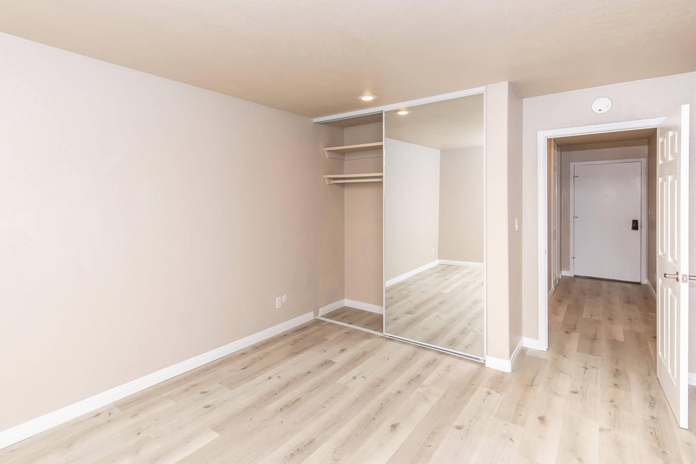 A spacious, empty bedroom featuring light beige walls and a wood laminate floor. There is a mirrored closet along one wall, and a doorway leading to a hallway is visible. The room has minimal decor, allowing for potential customization. Soft lighting enhances the airy atmosphere.