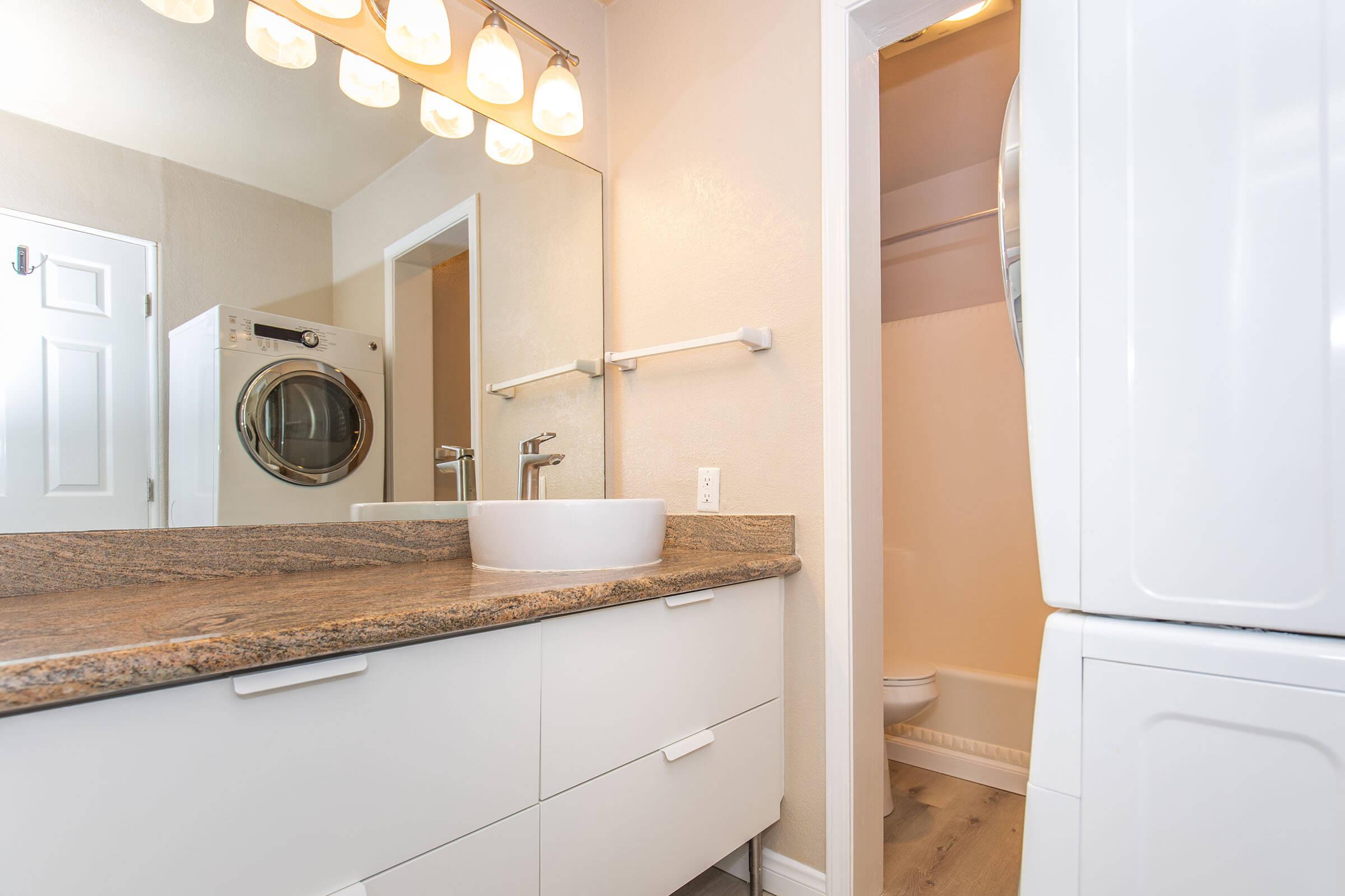 A modern bathroom featuring a vessel sink atop a granite countertop, with a large mirror and bright overhead lighting. A washer and dryer are visible in a closet area, and a small bathroom can be seen in the background, creating a functional and stylish space.