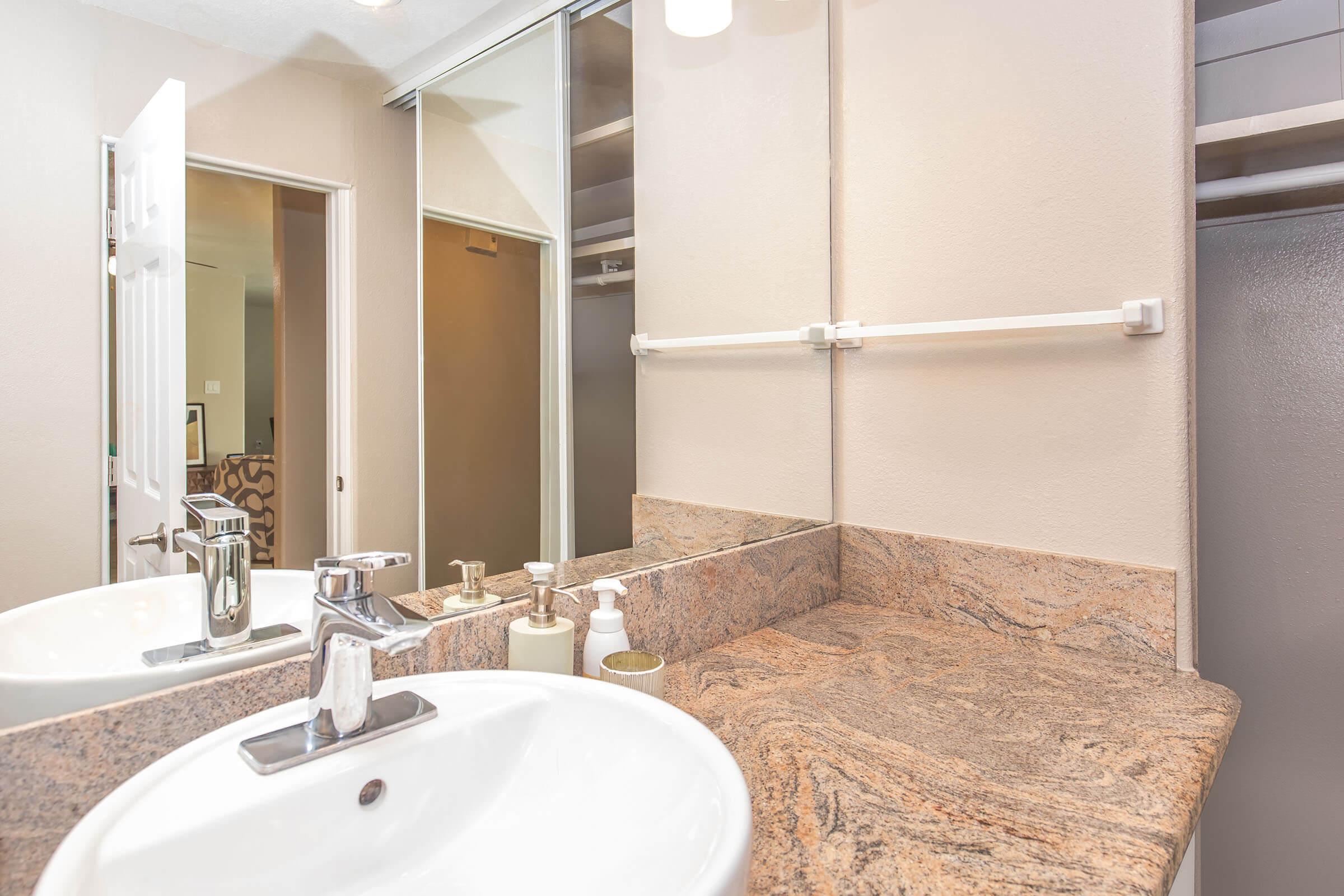 A clean bathroom countertop featuring a double sink with modern faucets, a granite surface, and decorative soap dispensers. A mirror reflects the space, and cabinets are visible in the background. The walls are painted in a neutral tone, contributing to a bright and airy atmosphere.